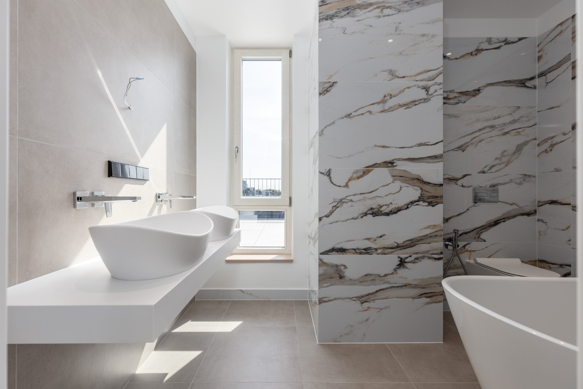 A modern bathroom featuring a freestanding white bathtub, wall-mounted faucets, and a window with a view of the outdoors. The room is decorated with large marble tiles on one wall and neutral-colored tiles on the floor and other walls, with natural l