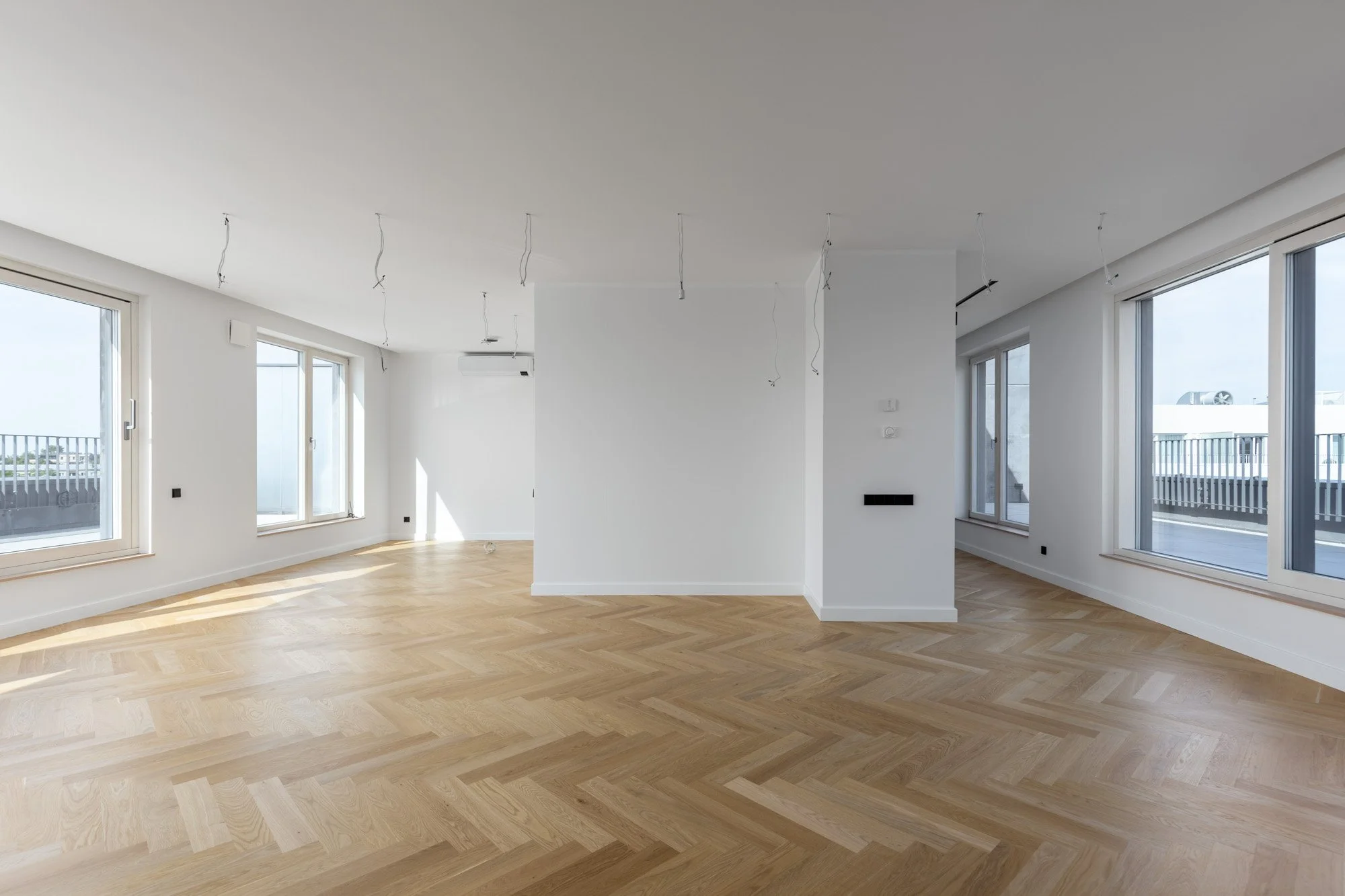 Empty room with large windows, light wood parquet floor, white walls, and ceiling with hanging wires indicating ongoing lighting installation.