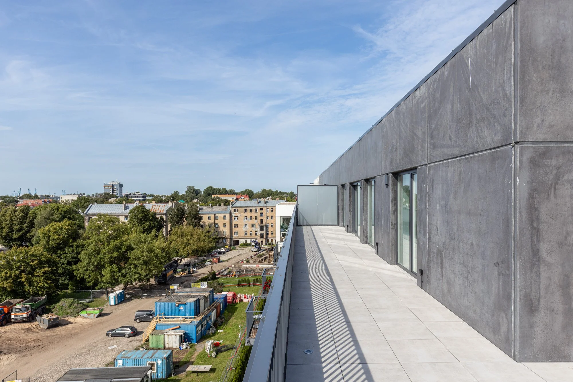 View of a modern building's rooftop terrace with a grey concrete exterior wall and glass doors, overlooking a city with trees, construction site, and other buildings under a partly cloudy sky.