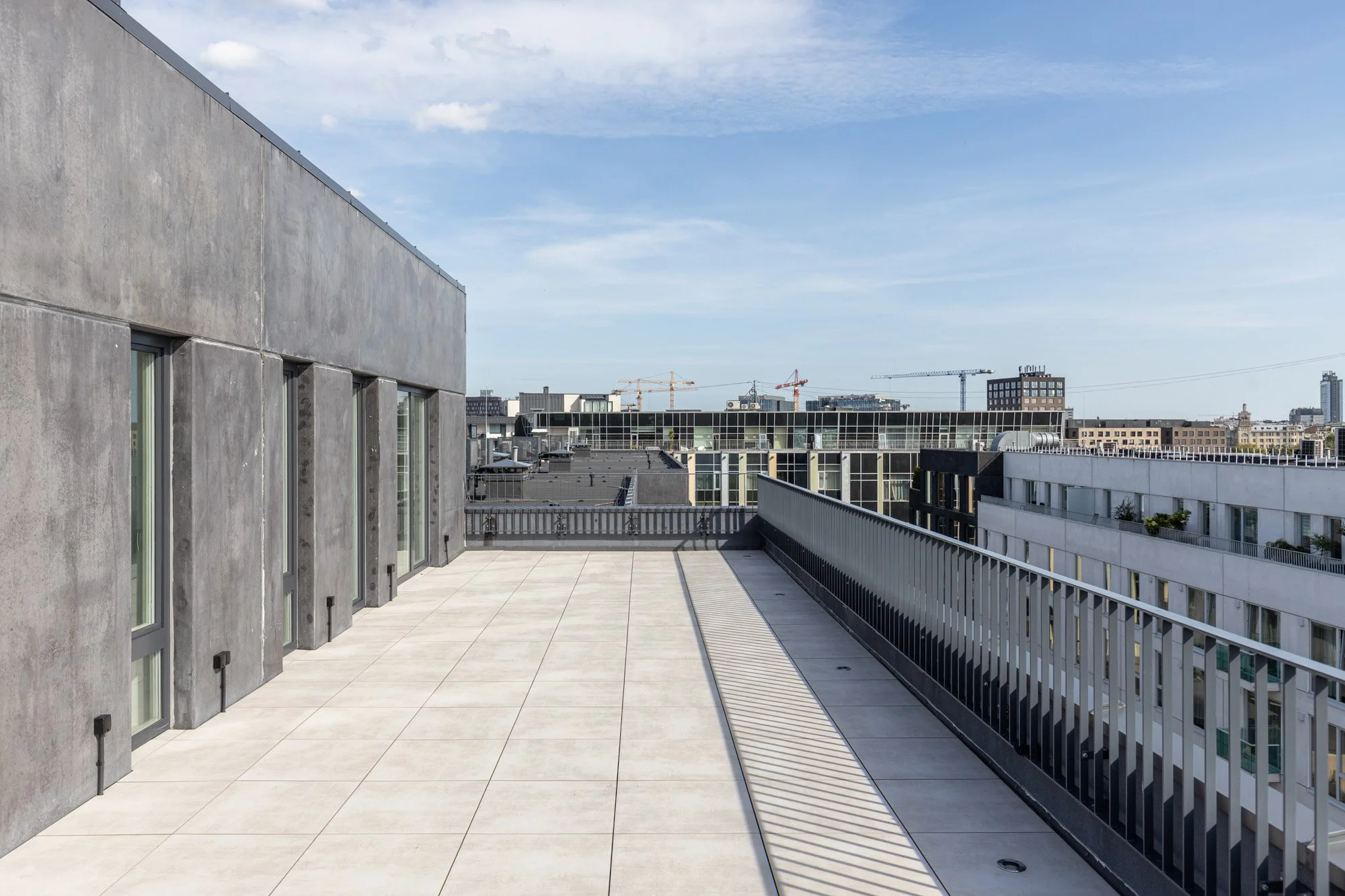 Cityscape view from a modern rooftop terrace with concrete flooring and metal railing, featuring high-rise buildings and construction cranes under a partly cloudy sky.