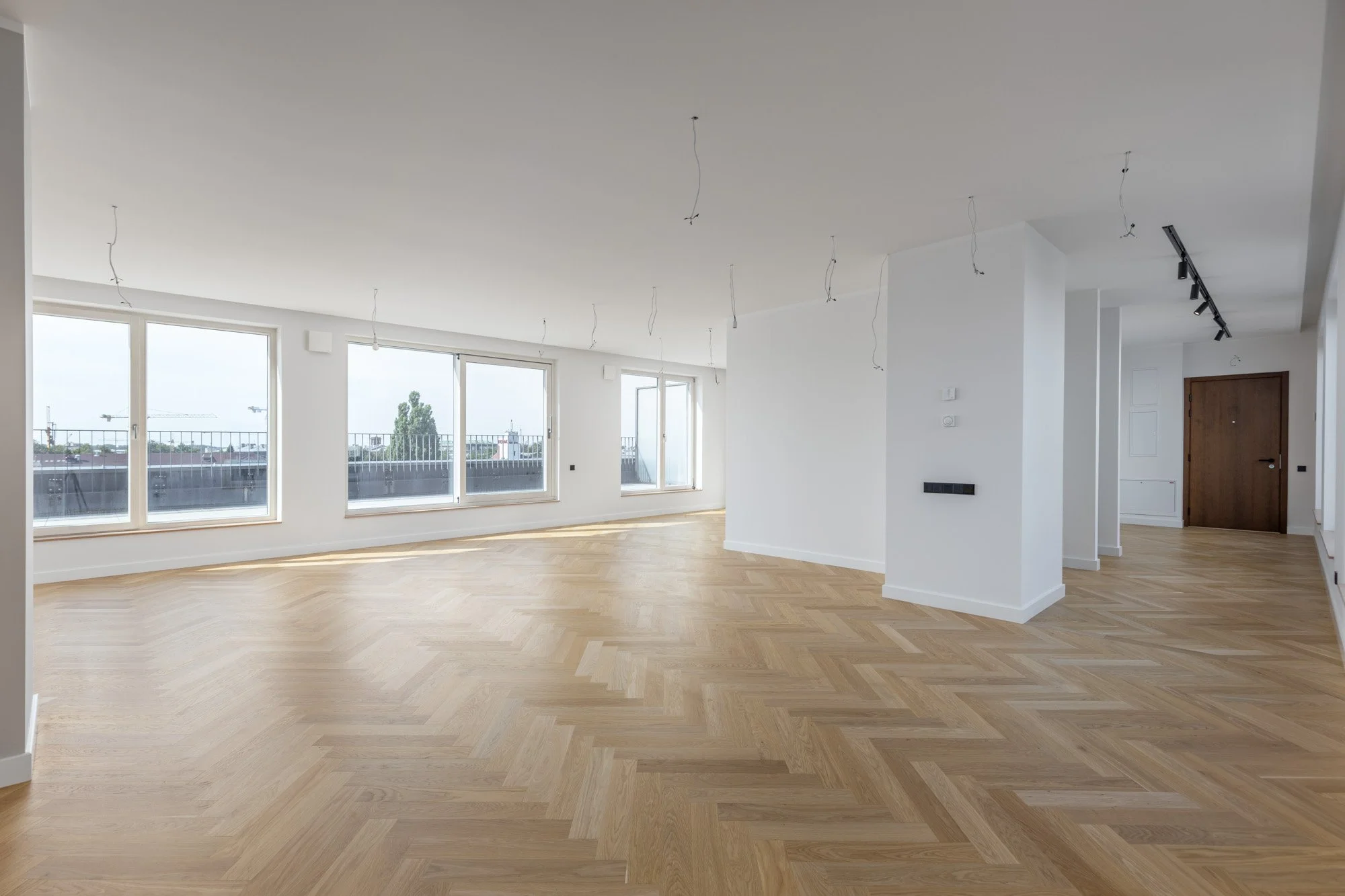 Empty modern living room or apartment with large windows and light wooden flooring, showing walls with wiring and track lighting installation in progress.