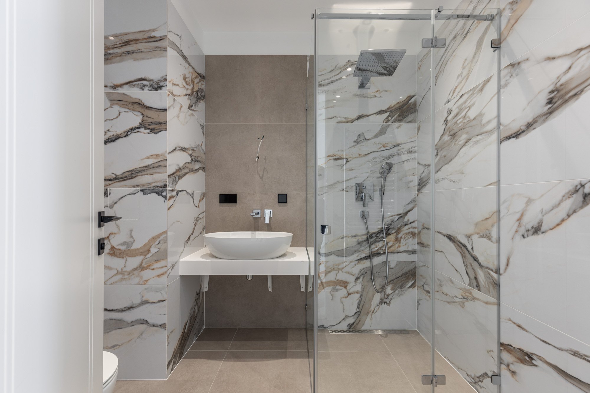 Modern bathroom with marble walls, a glass-enclosed shower with a rainfall showerhead and handheld shower, a white vessel sink, and a beige wall with electrical outlets.