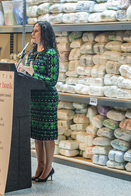 Executive Director Beverly Hamilton, standing at podium, speaking at Austin Diaper Bank's warehouse grand opening with a huge shelf of diapers in the background.