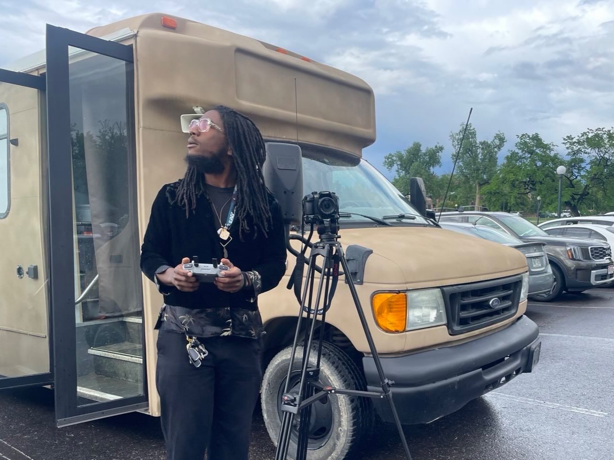 A man with dreadlocks, wearing sunglasses and a black jacket, holding a drone controller, standing next to a beige RV with a camera mounted on a tripod. The scene is in a parking lot with trees and other vehicles in the background.