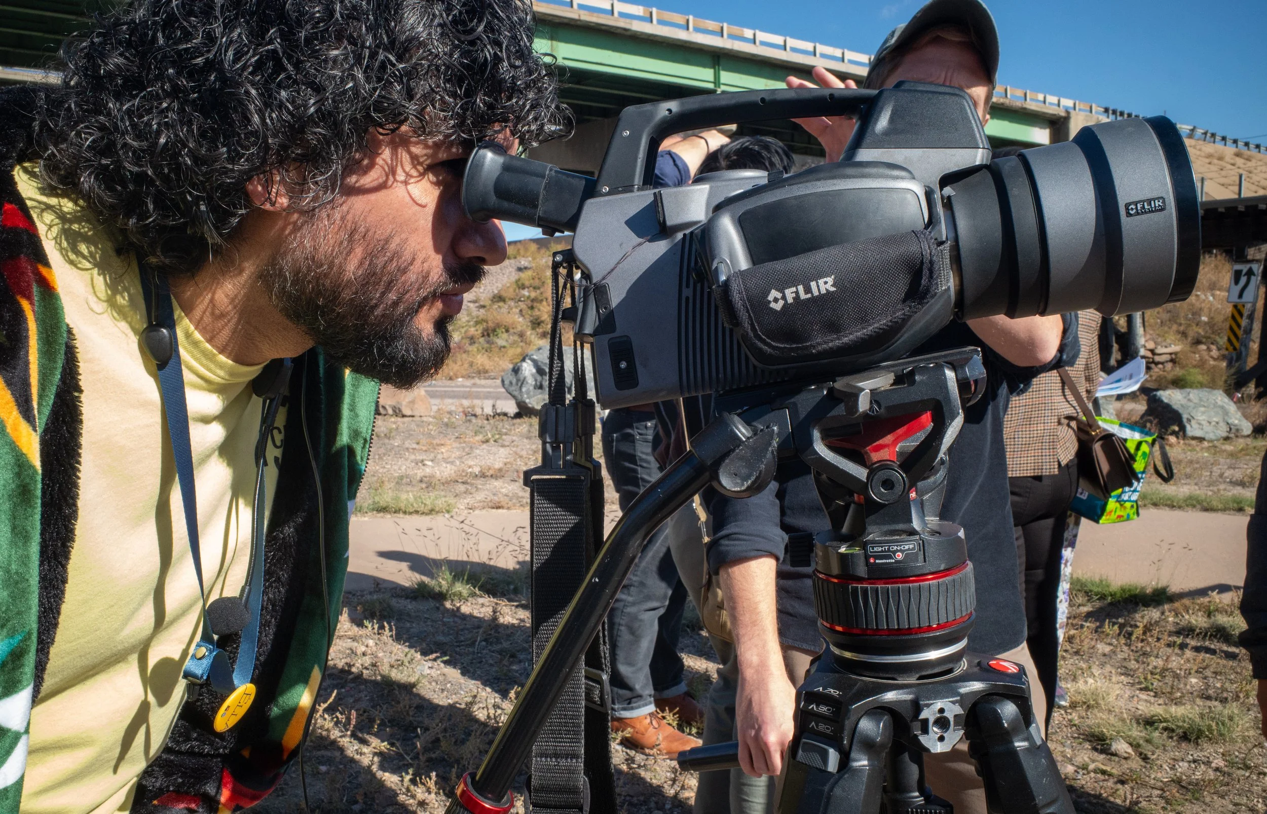 Group of people working with a professional video camera outdoors near a bridge in sunny weather.