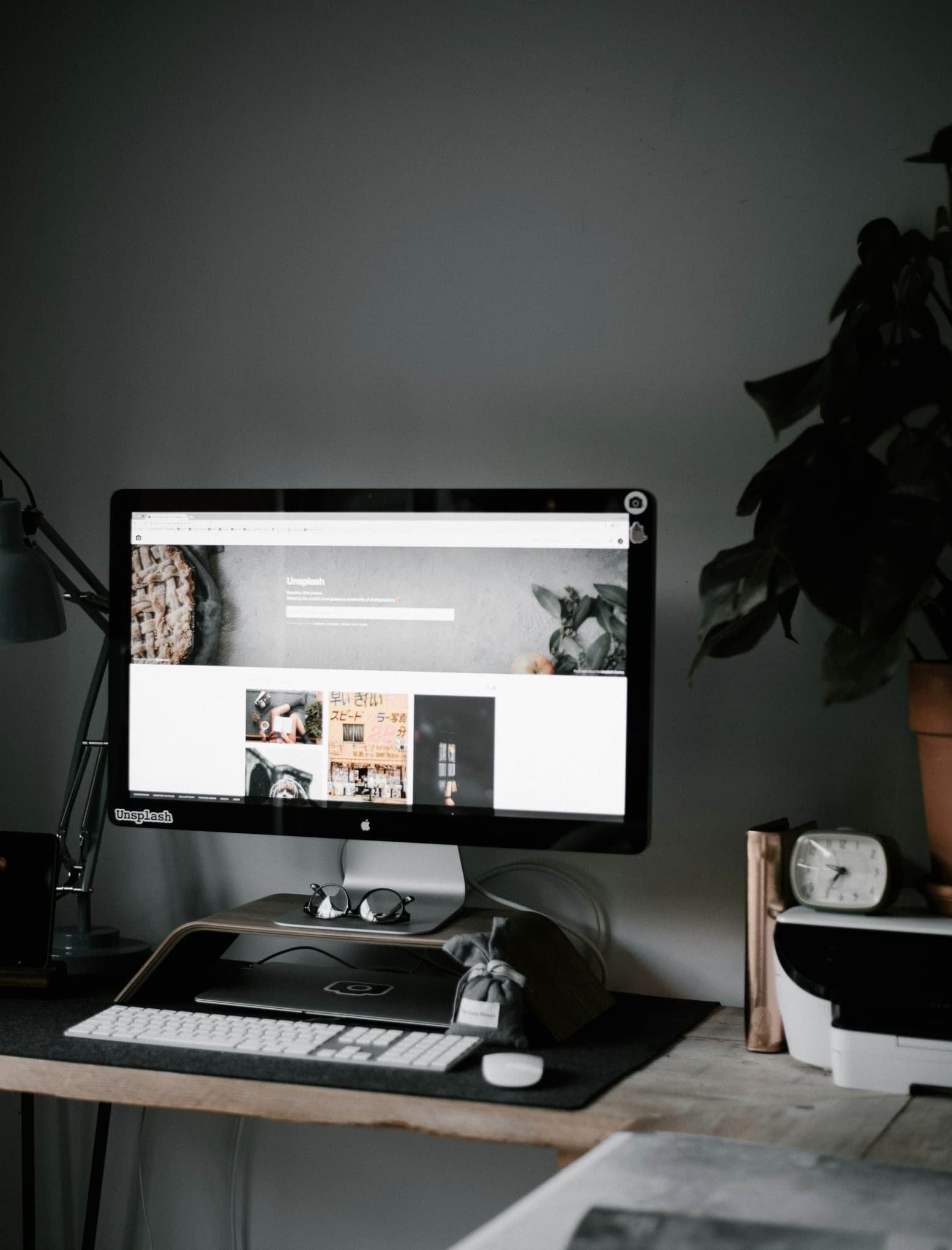 A workspace with an iMac on a wooden desk, showing a webpage, with a keyboard, mouse, pair of glasses, and a small potted plant nearby.