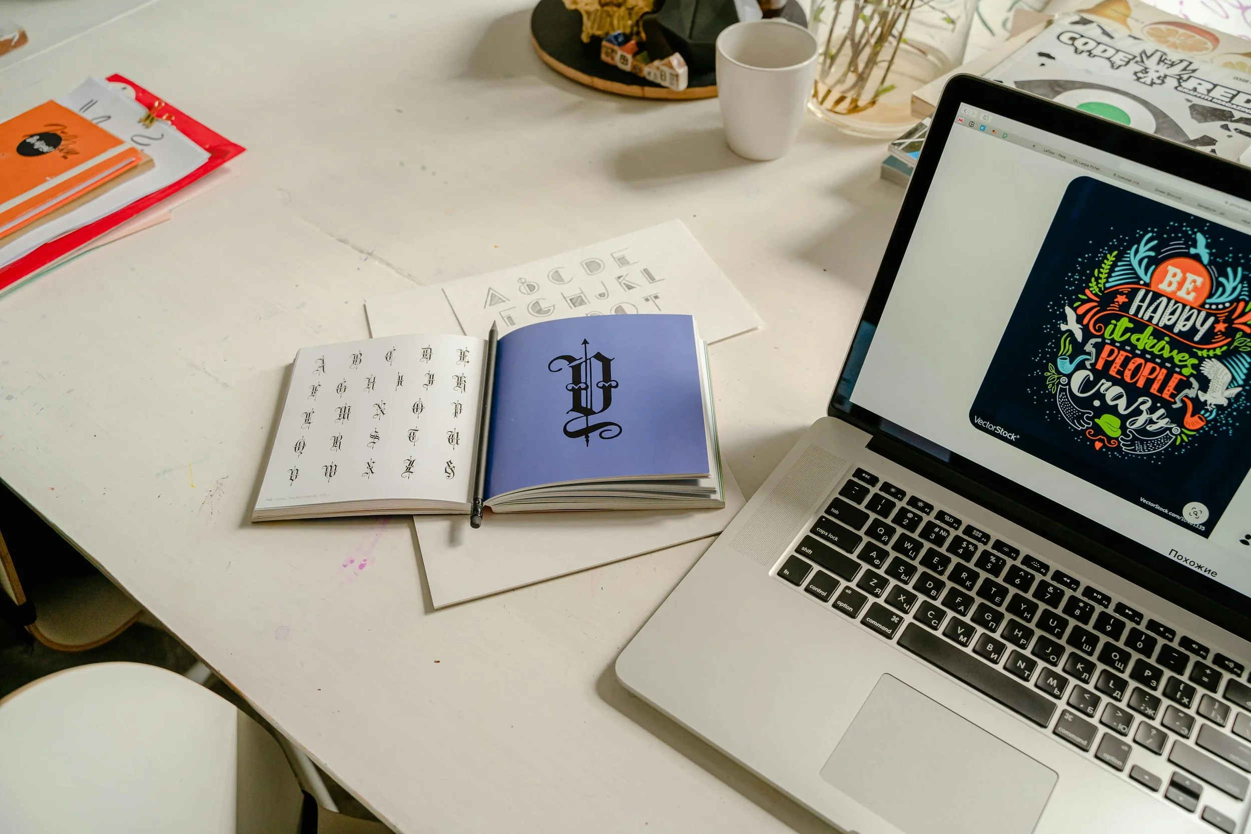 A white desk with various books, a laptop displaying a colorful graphic with the text 'Be happy! It drives people crazy,' a white mug, and some papers and notebooks scattered around.