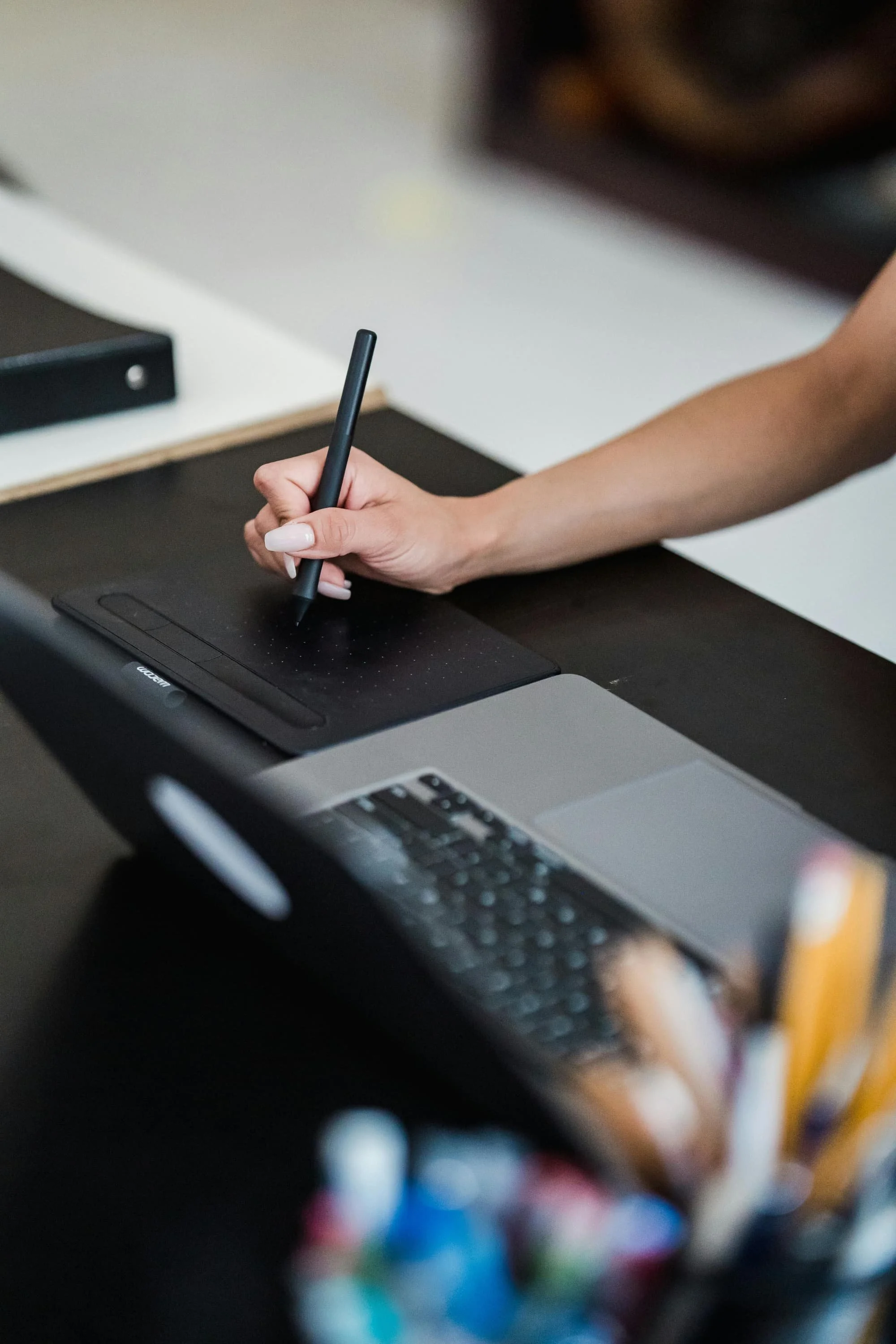 A person using a black stylus tablet on a desk, with a laptop and office supplies visible.
