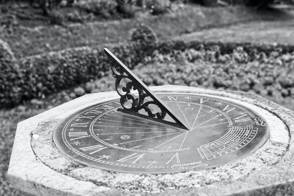 A sundial with a decorative gnomon in an outdoor garden setting, showing the time with shadows, in black and white.