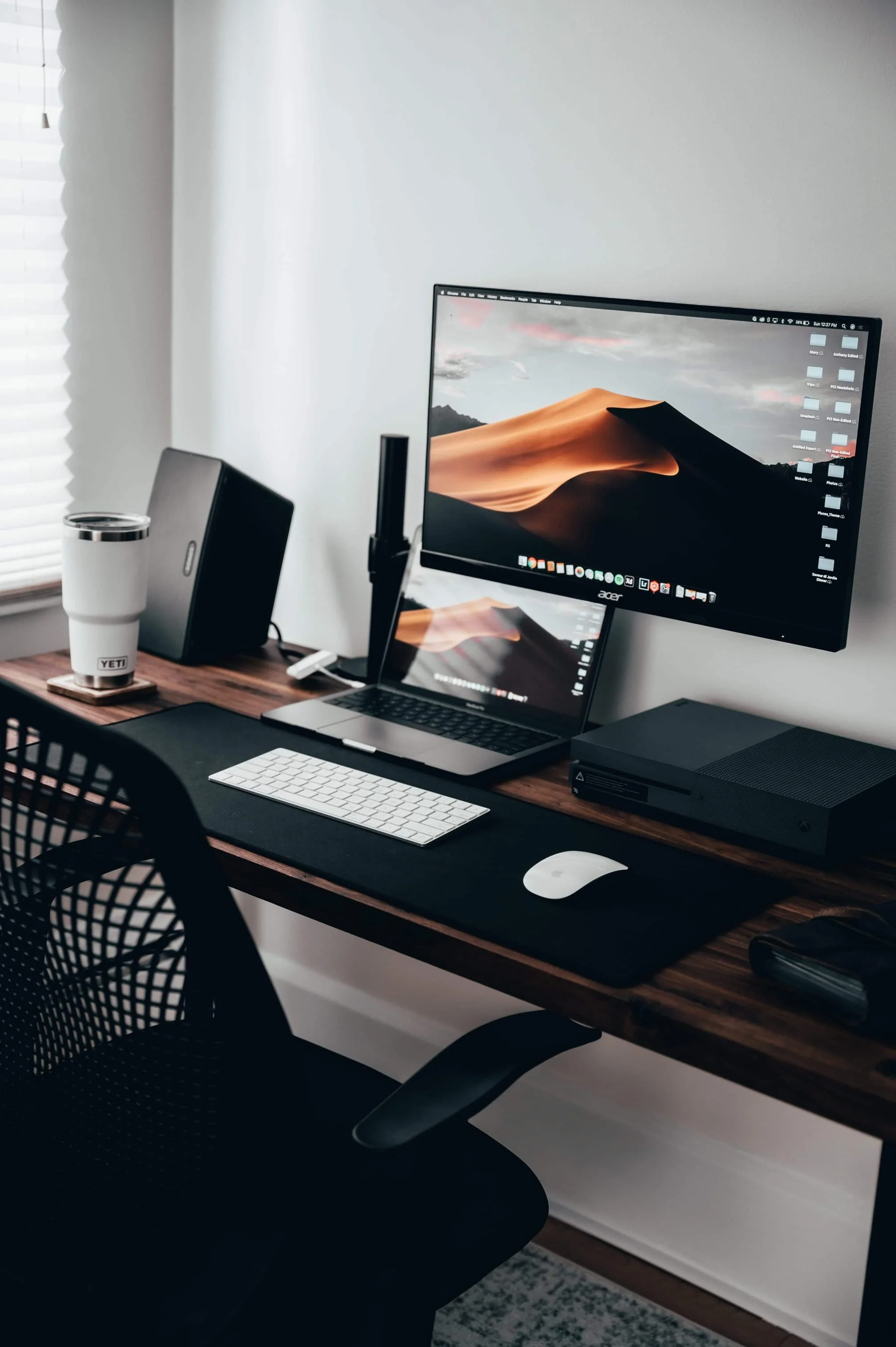 A modern home office setup with a large computer monitor displaying a desert landscape wallpaper, a laptop, a white keyboard, and a white mouse on a black desk mat, a desktop tower, a speaker, a YETI tumbler, and a black mesh office chair in front of a window with blinds.
