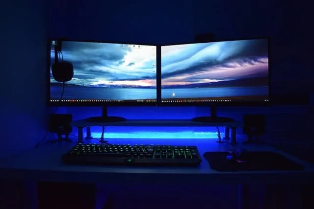 Dual curved computer monitors displaying a scenic sky and water landscape in a dark room with blue LED lighting, a keyboard, mouse, and speakers on the desk.