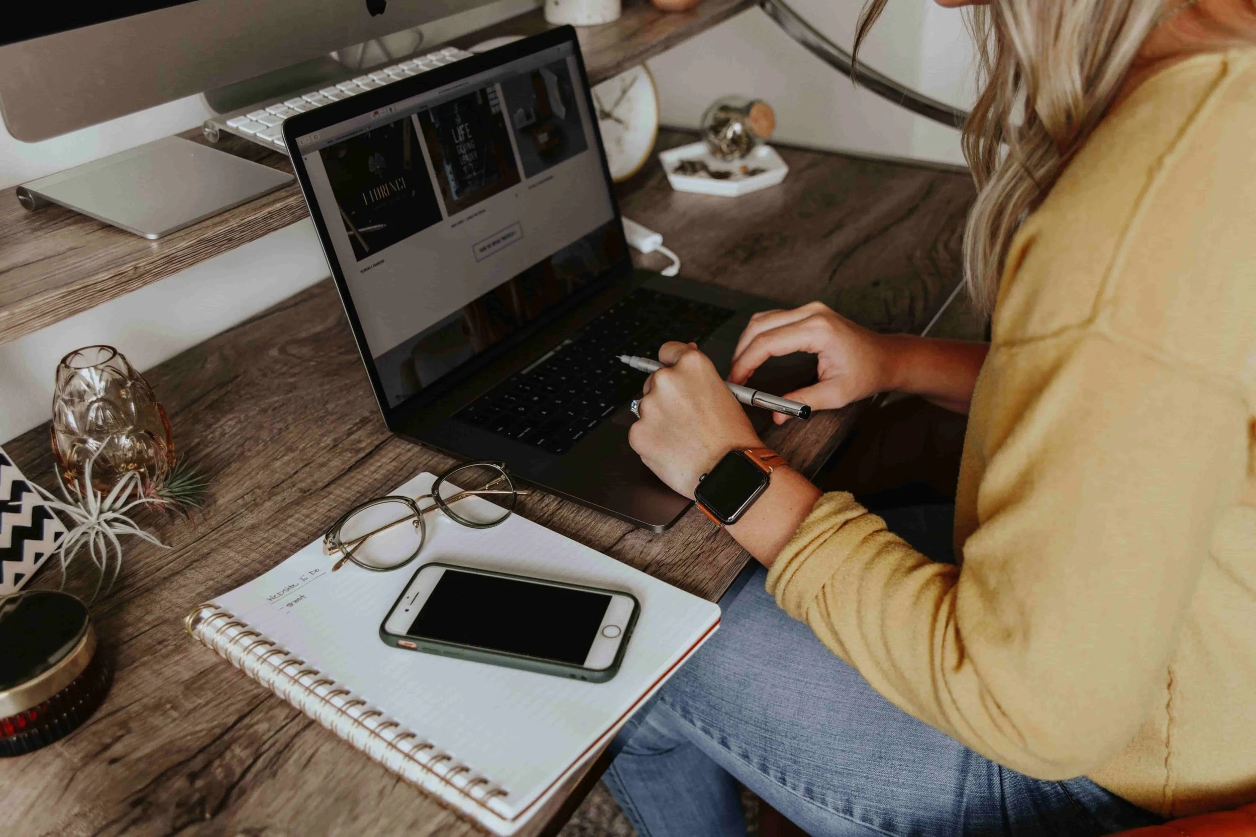A woman in a yellow shirt sitting at a wooden desk, working on a laptop, with glasses, a smartphone, a notebook, and other office supplies on the desk.