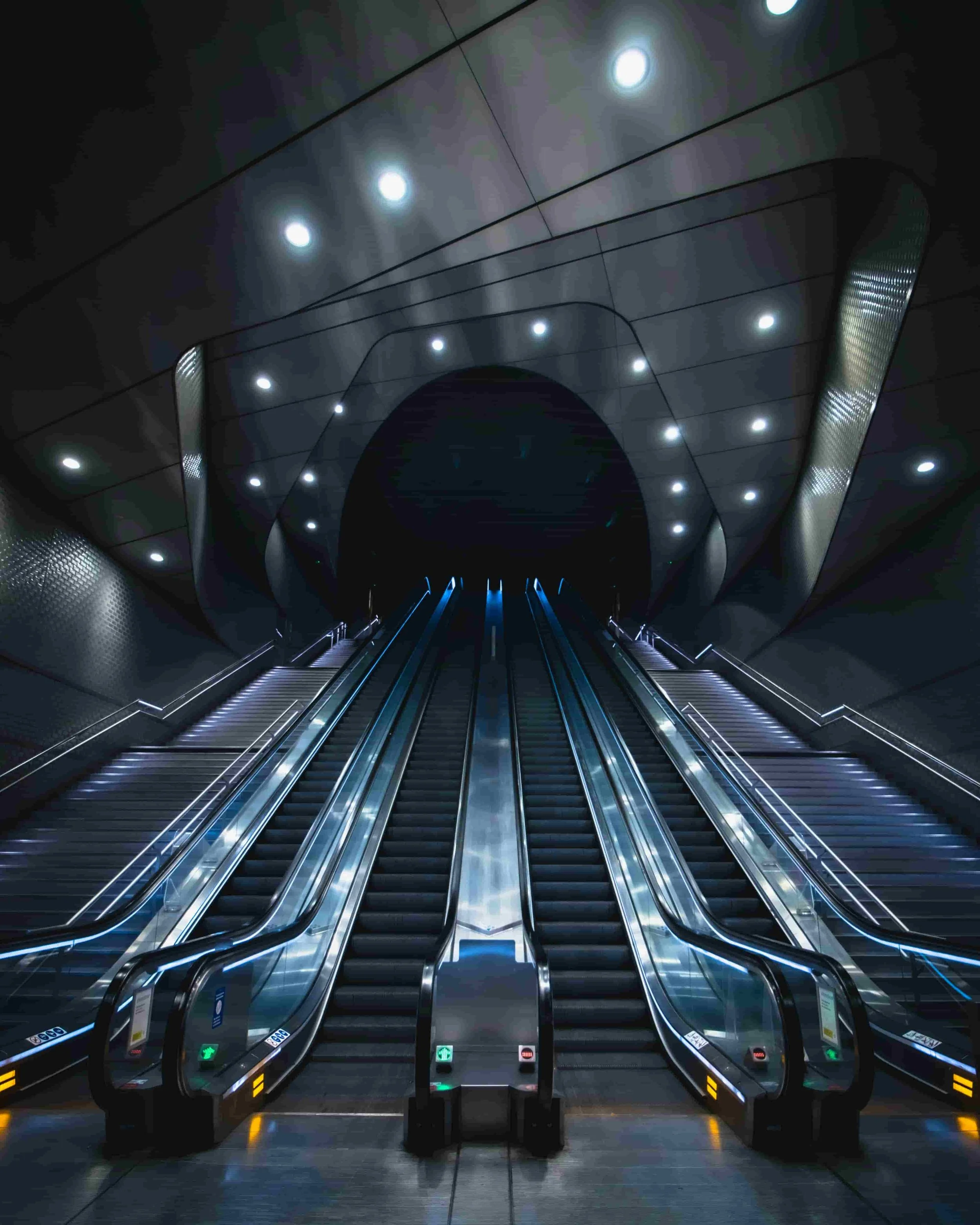 Empty escalators in a modern, dark underground subway or metro station with a sleek, futuristic design and bright overhead lighting.