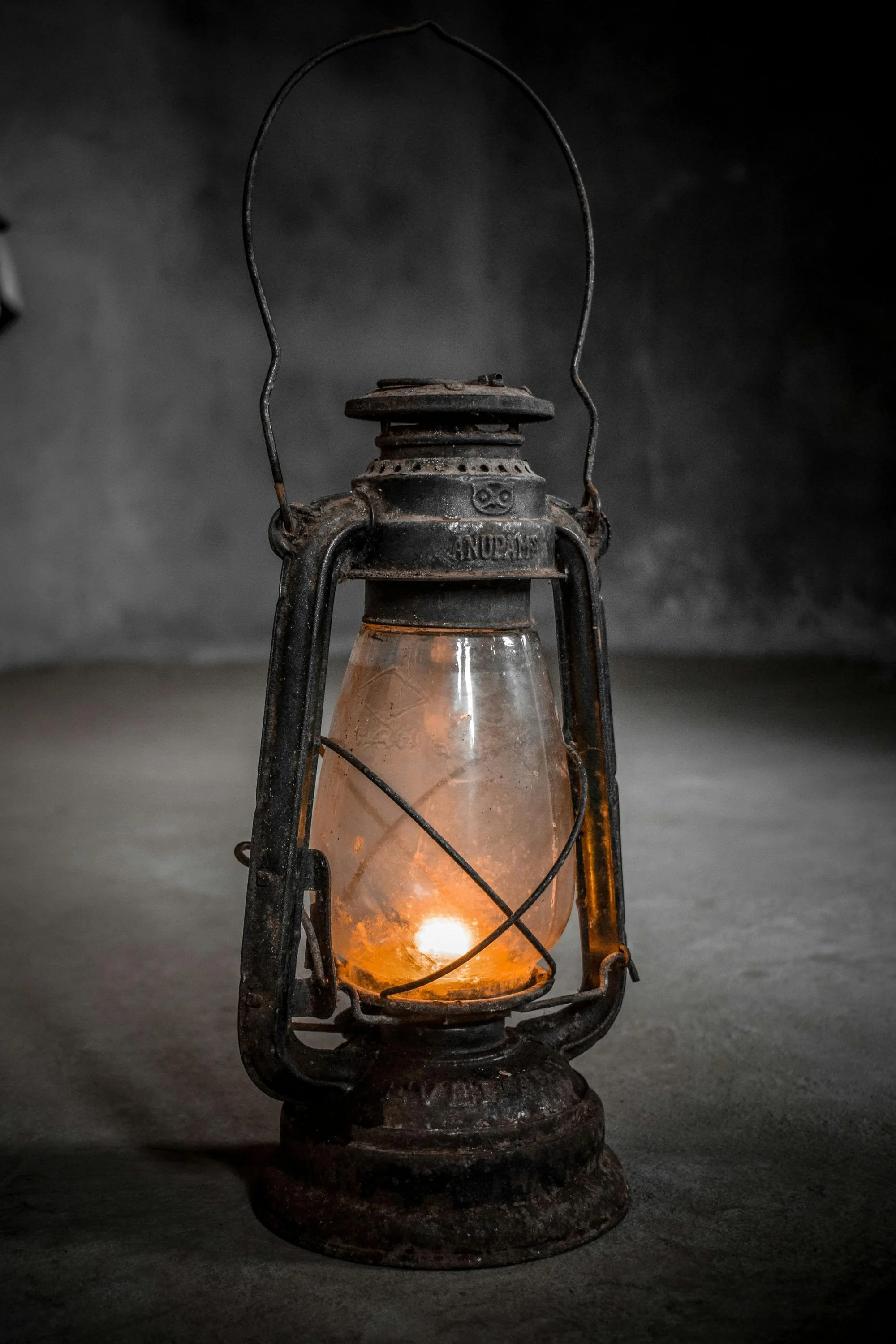 An old, rusty kerosene lantern with a glowing flame inside, sitting on a concrete surface against a dark, blurred background.