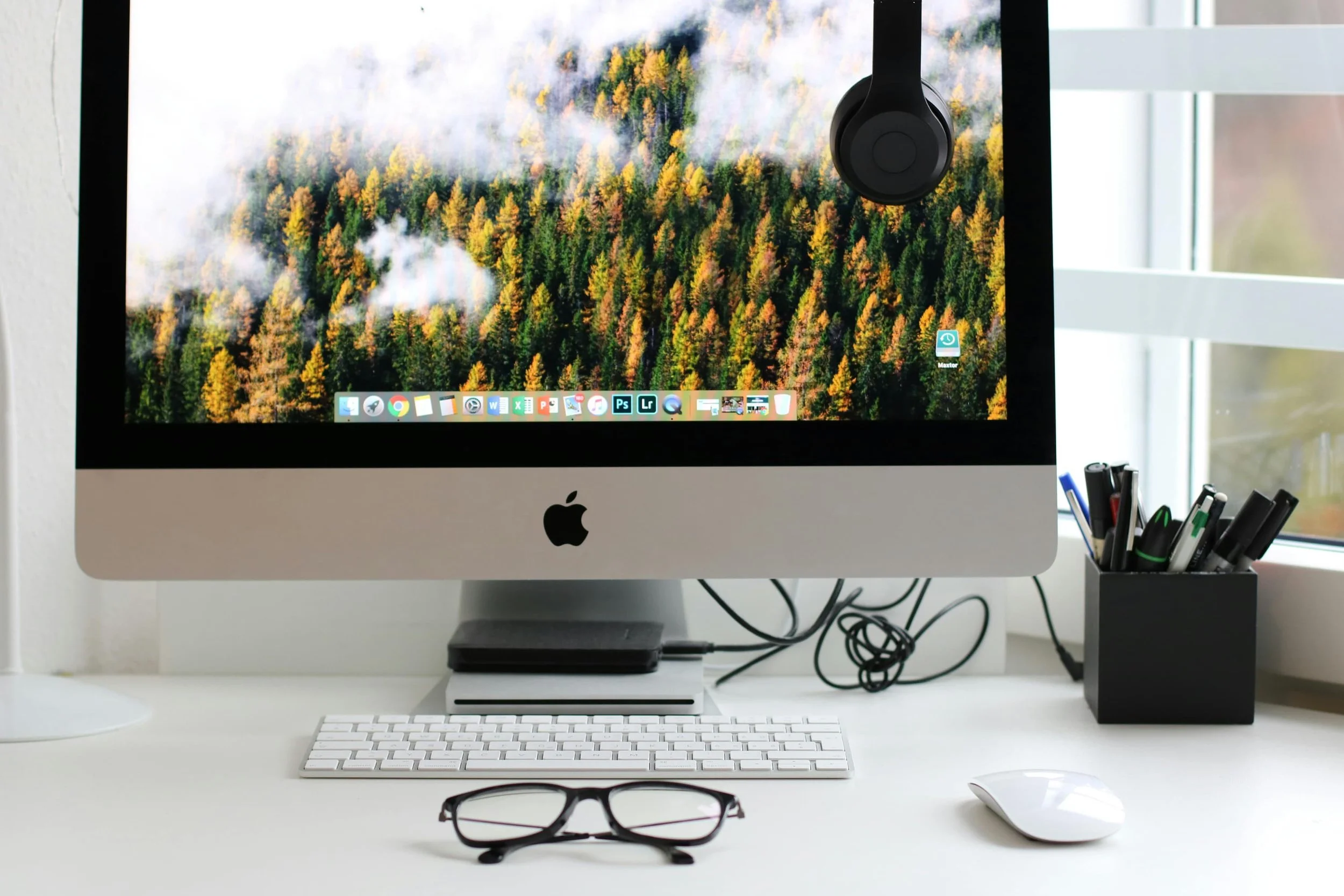 An iMac computer on a white desk with a small white keyboard, white mouse, and black eyeglasses in front. The screen shows a forest landscape with trees and clouds. To the right, a black pen holder with pens and markers, and a window with blinds in the background.