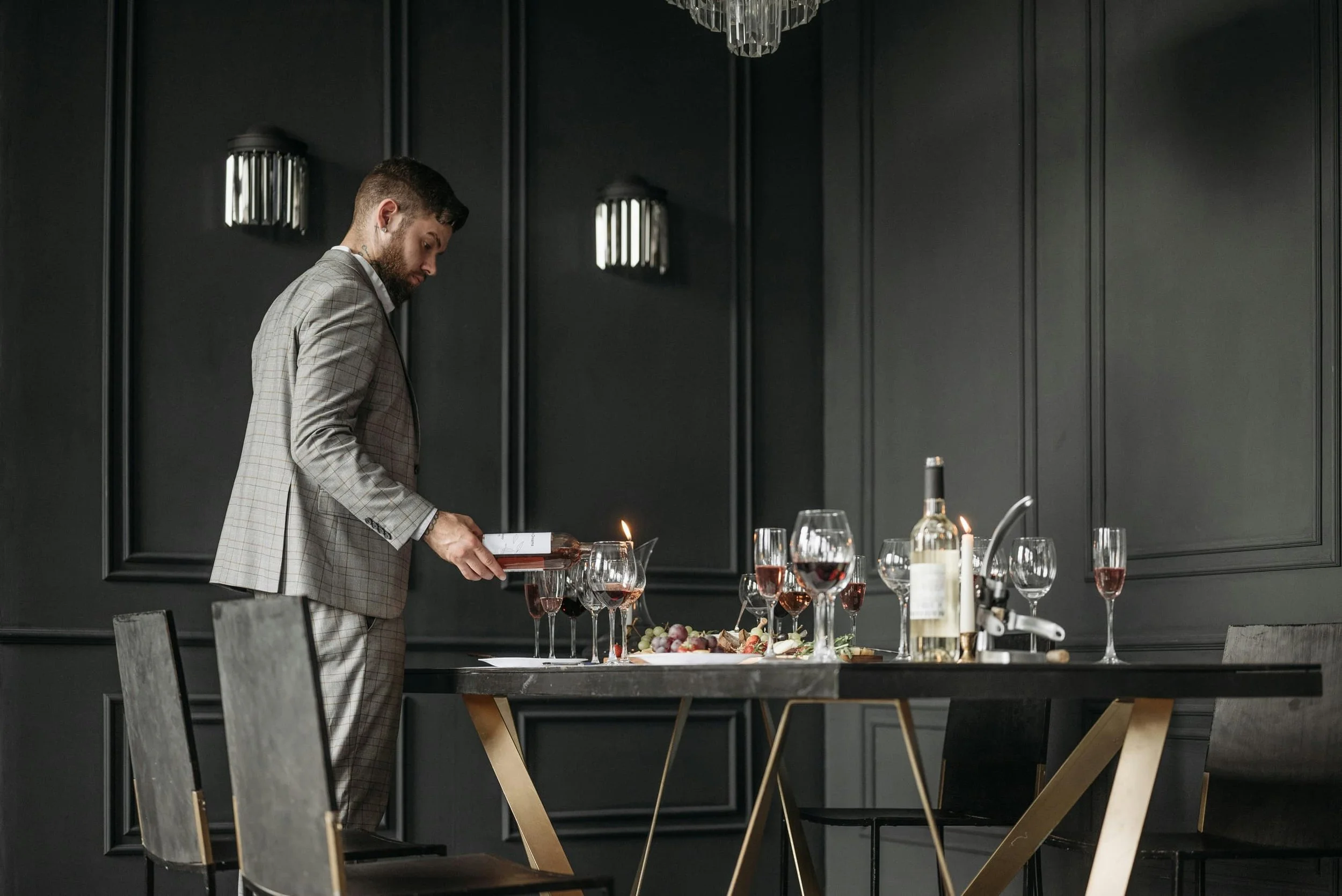 A man in a checkered suit pours red wine into glasses on a table set for a meal in a dark, elegant dining room.