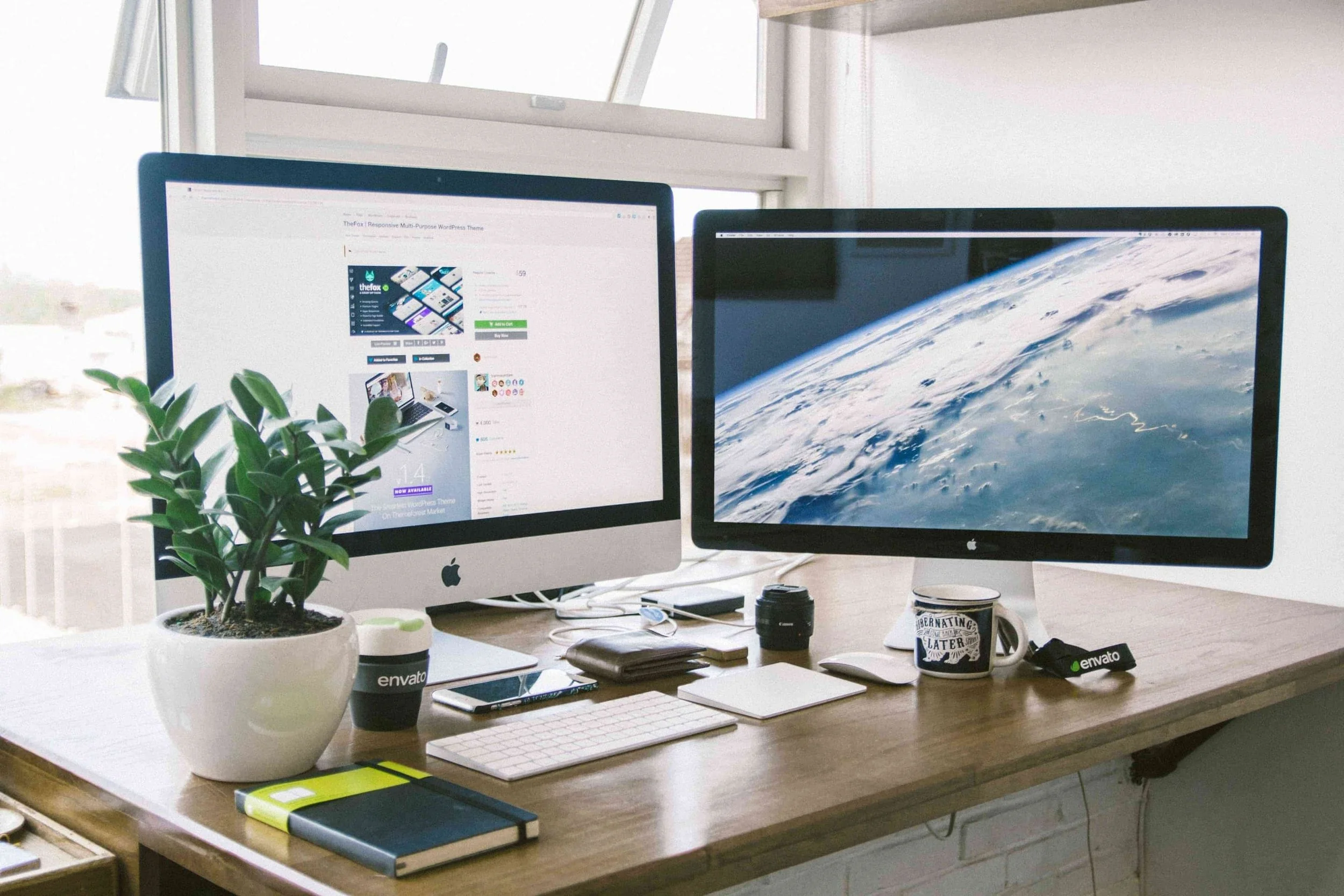 Desk with two Apple monitors, a potted plant, notebooks, a smartphone, a mug, a camera lens, a keyboard, a mouse, and an audio device with the Envato logo, in front of a window showing an urban landscape.