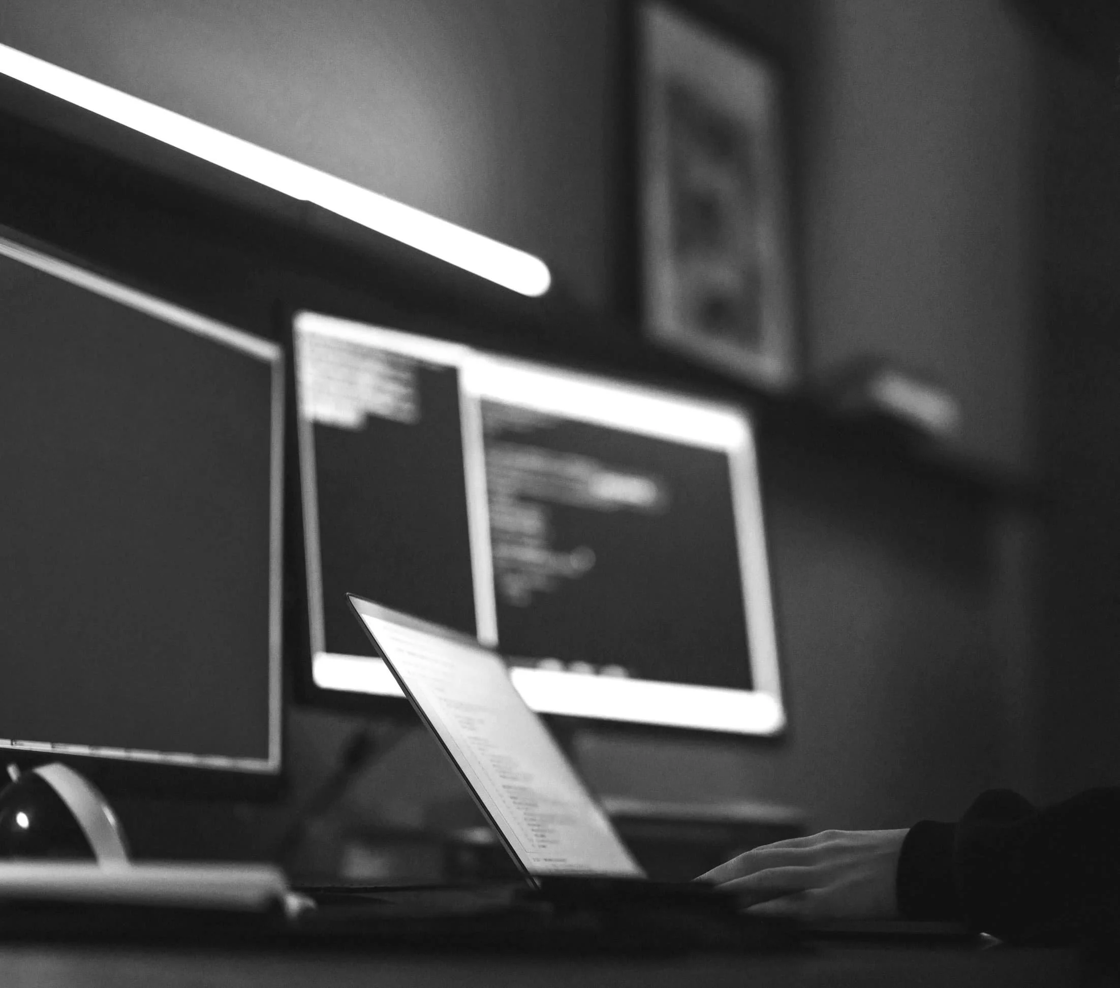 Black and white photo of a person's hand typing on a laptop, with two monitors and a tablet displaying code on a desk.