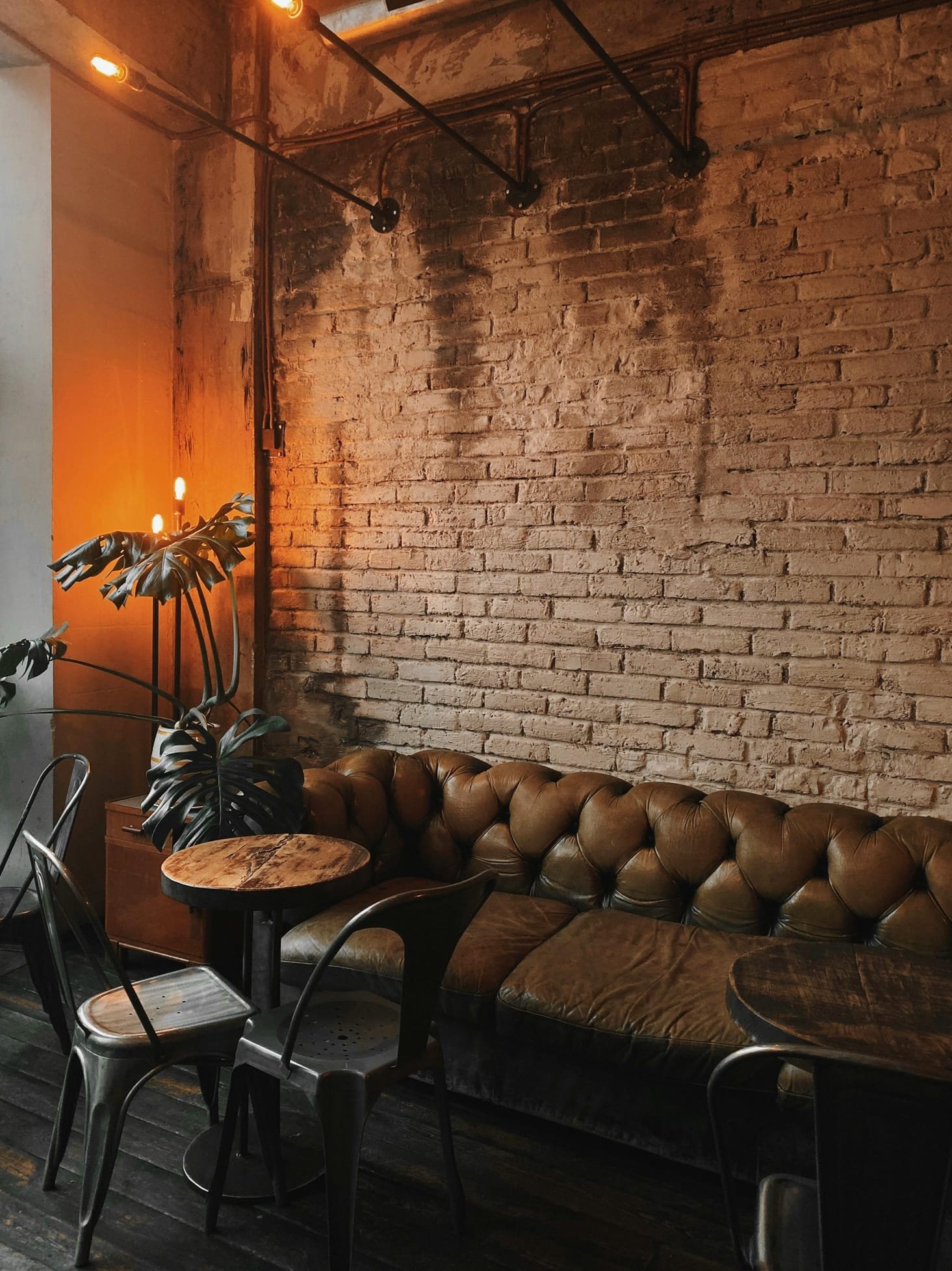 Interior of a cozy cafe with exposed brick walls, a green leather tufted sofa, a small round wooden table, metal chairs, and warm hanging light bulbs creating a dim, inviting atmosphere.