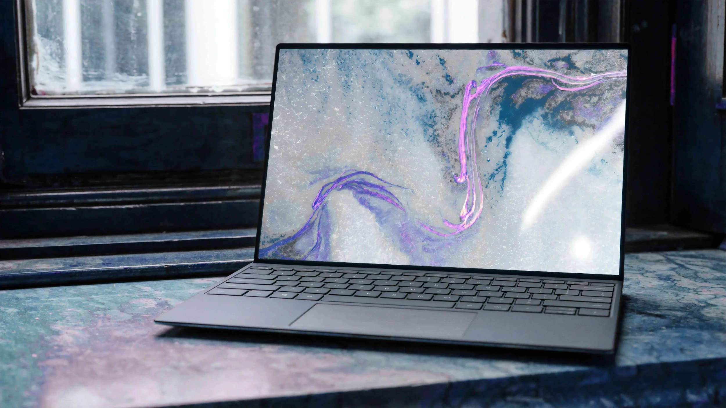 A sleek, modern laptop with a thin design, open on a windowsill with a textured, colorful surface. The screen displays an abstract artwork with swirling purple and blue lines, set against a light, speckled background. The window behind the laptop shows a blurred outdoor scene, allowing natural light to illuminate the workspace.