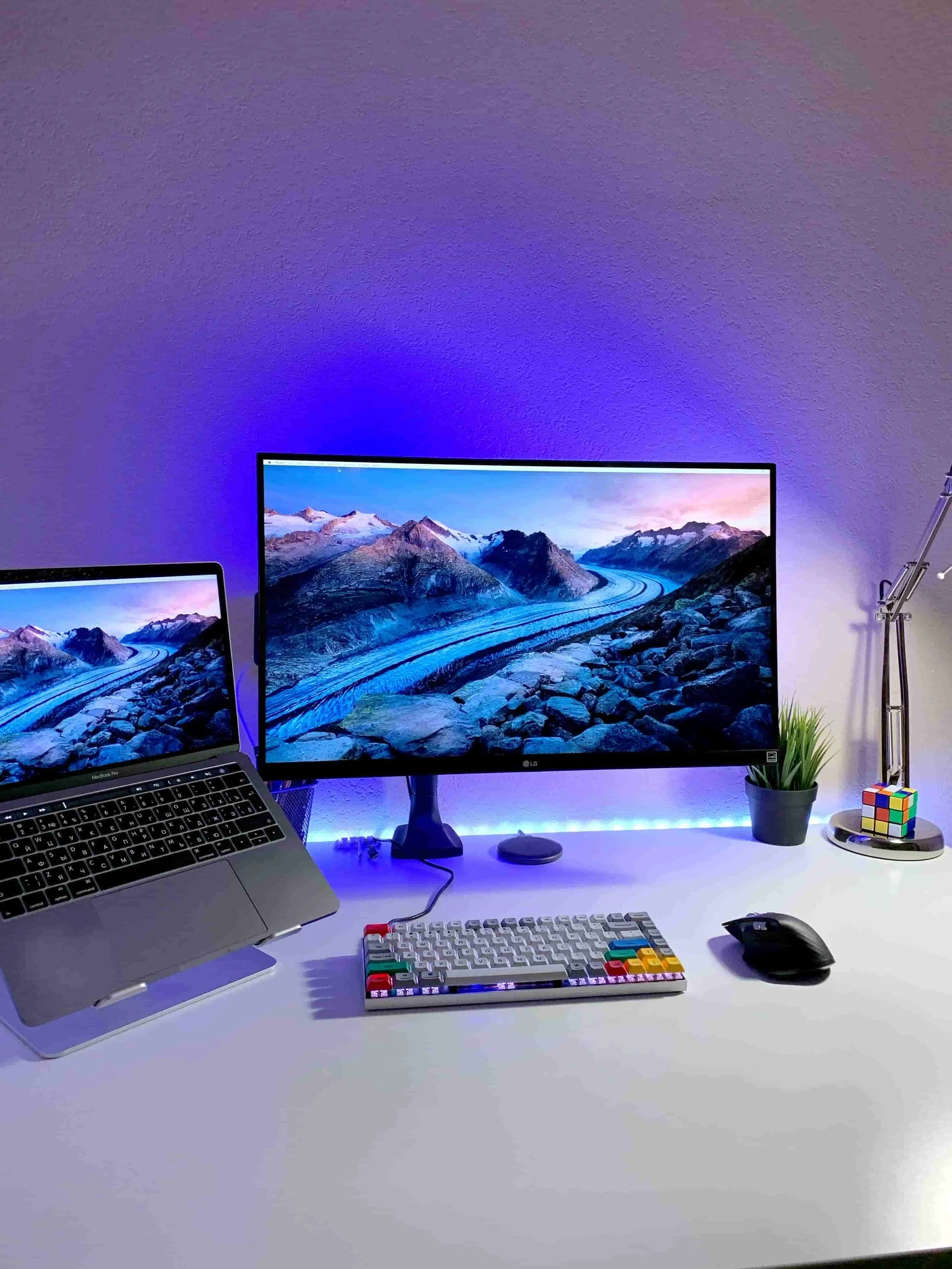 A minimalist workspace with a white desk, two monitors displaying mountain landscapes, a laptop on a stand, a mechanical keyboard with colorful keycaps, a black computer mouse, a small potted plant, a Rubik's cube on a round stand, and a modern adjustable desk lamp, illuminated by purple and blue LED lighting on the wall.