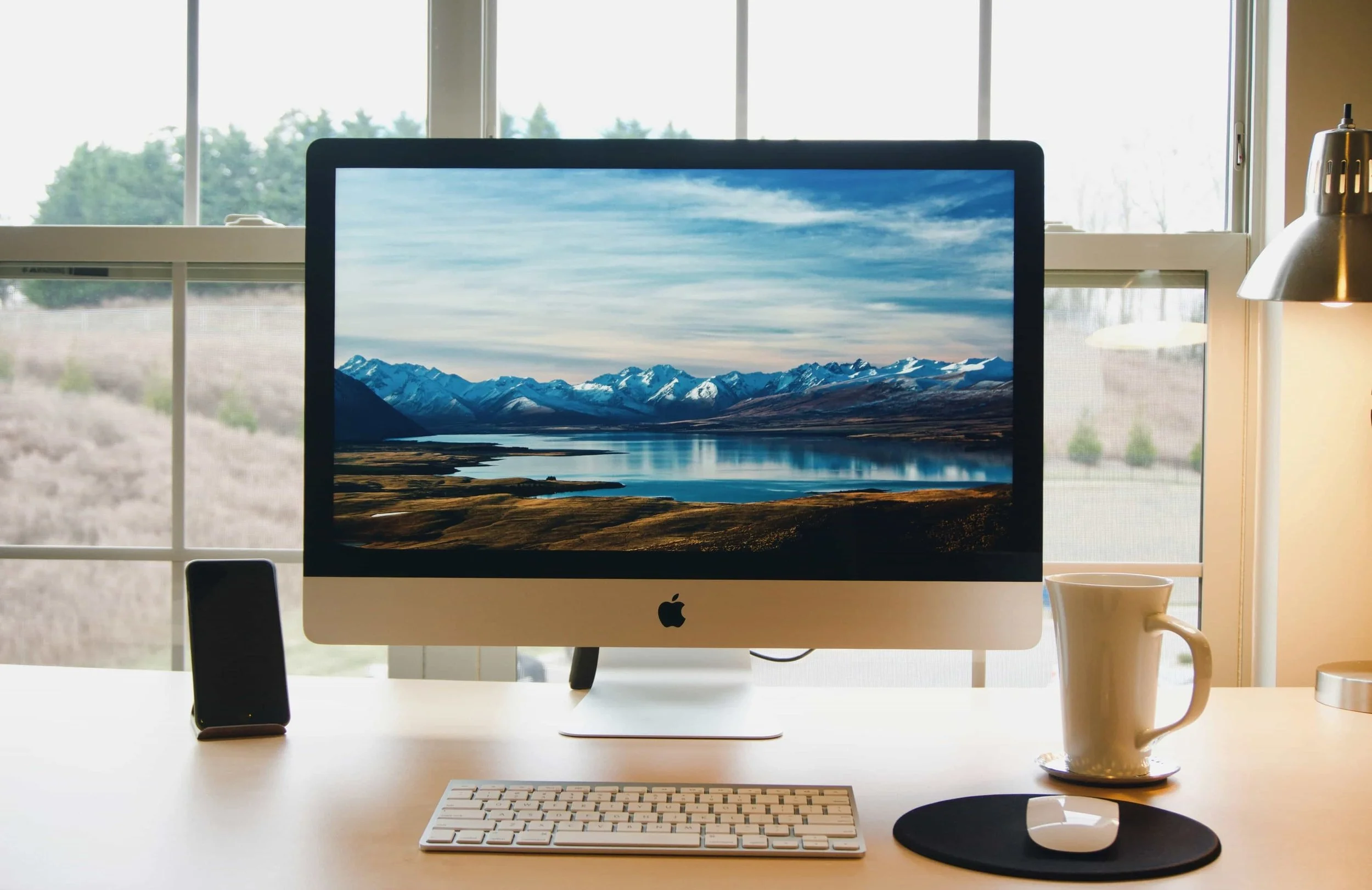 Desk with an Apple iMac computer displaying a mountain lake landscape, a white keyboard, a white mouse on a black mouse pad, a coffee mug, a smartphone, and a desk lamp in front of a window showing an outdoor scene.