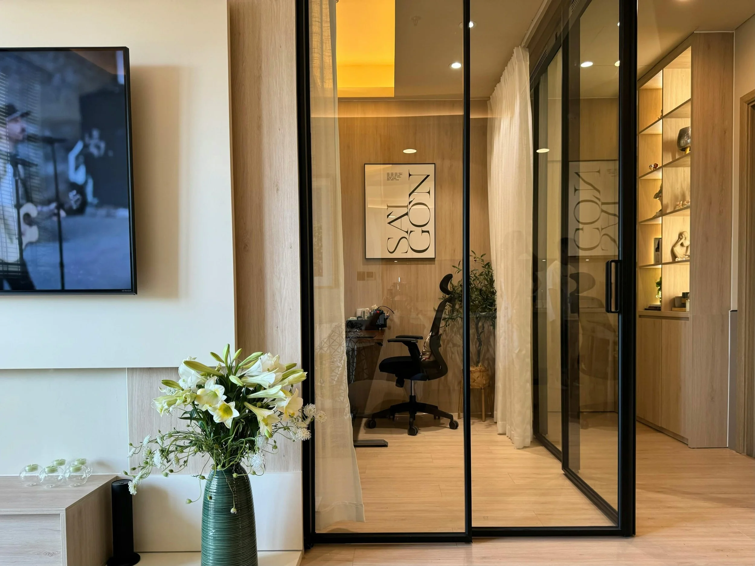 Modern interior room with a glass partition, a black office chair behind a desk, a large framed artwork on the wall with the text 'SAY GONE', and a wooden shelf with decorative items.
