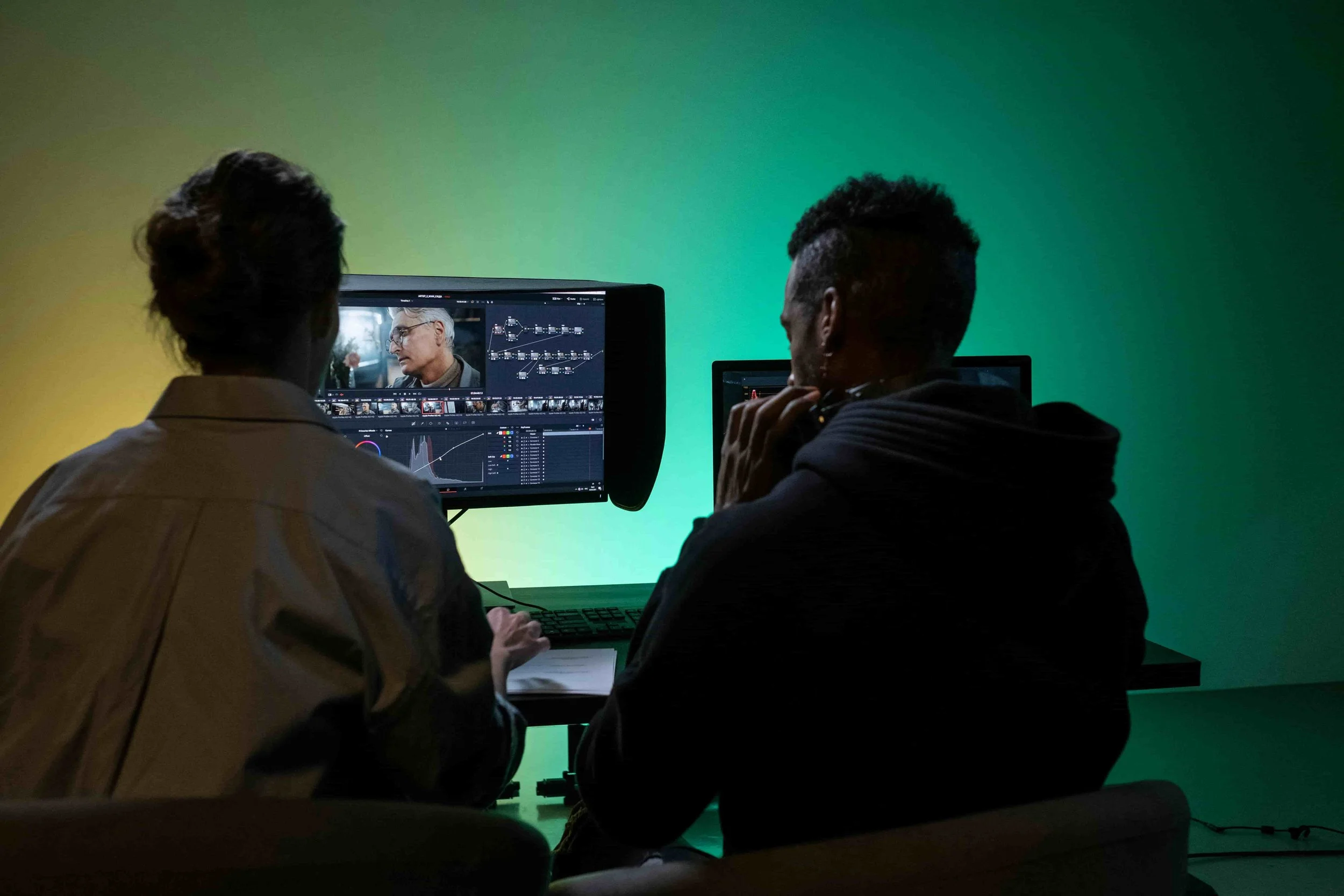 Two people working on a film editing project in a dark room with a green screen background, watching footage on a monitor.