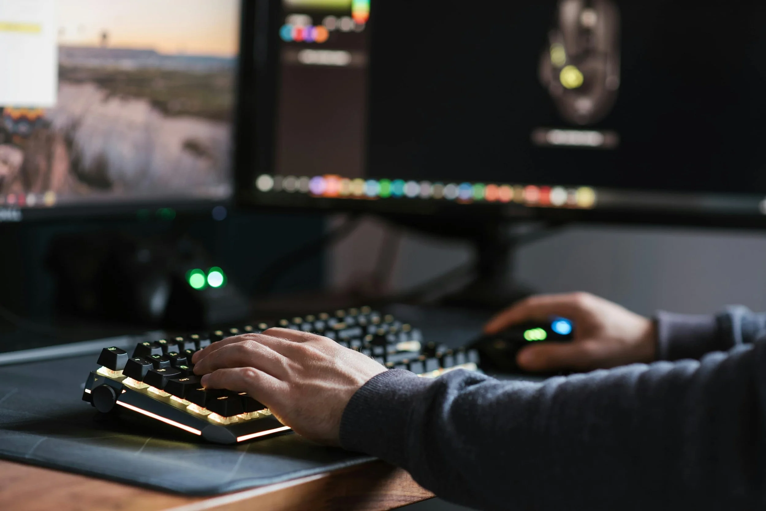 Person using a computer with dual monitors, a mechanical keyboard with backlit keys, and a gaming mouse in a dark room.