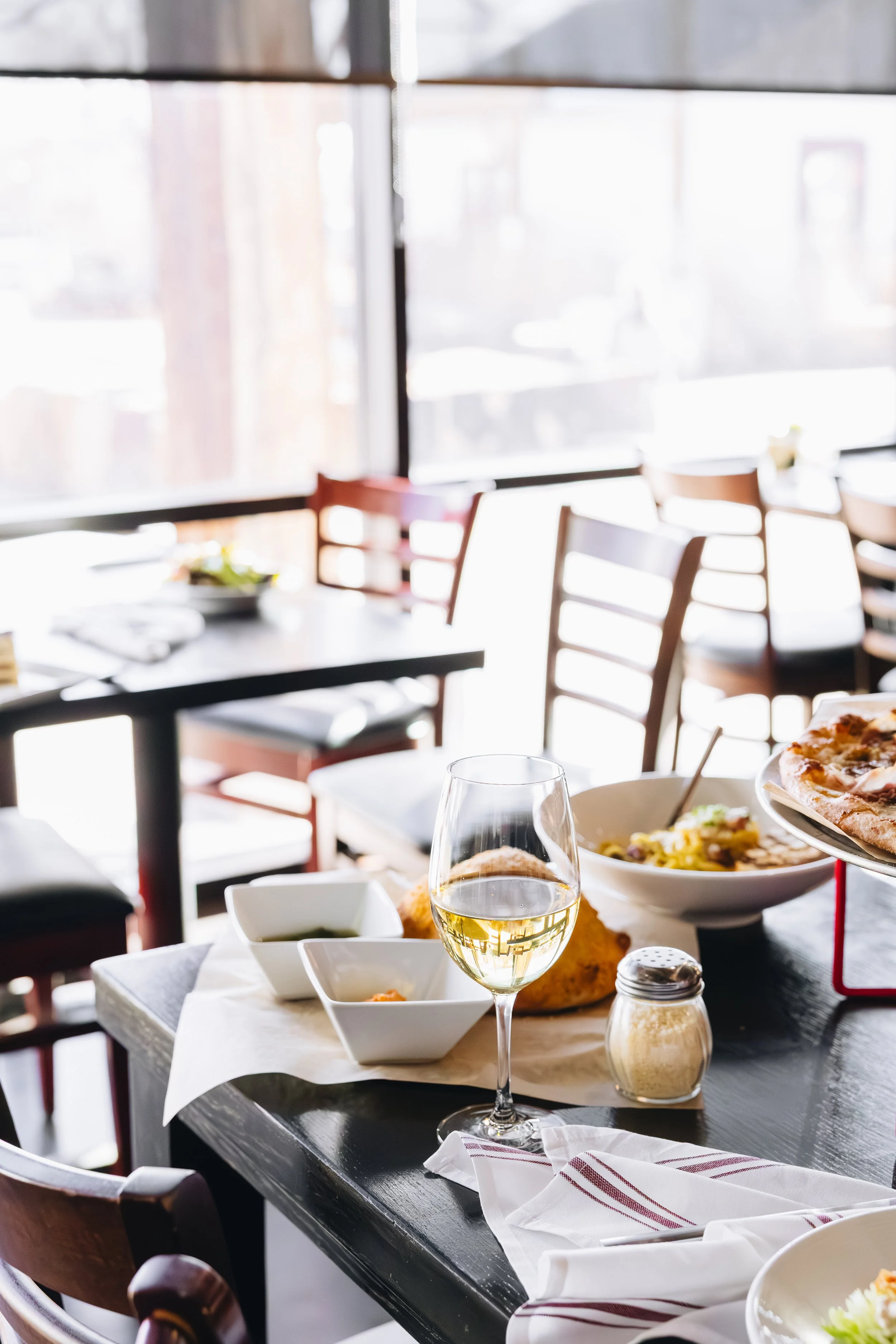 Restaurant table with a glass of white wine, pizza, pasta, dips, and a cheese shaker.