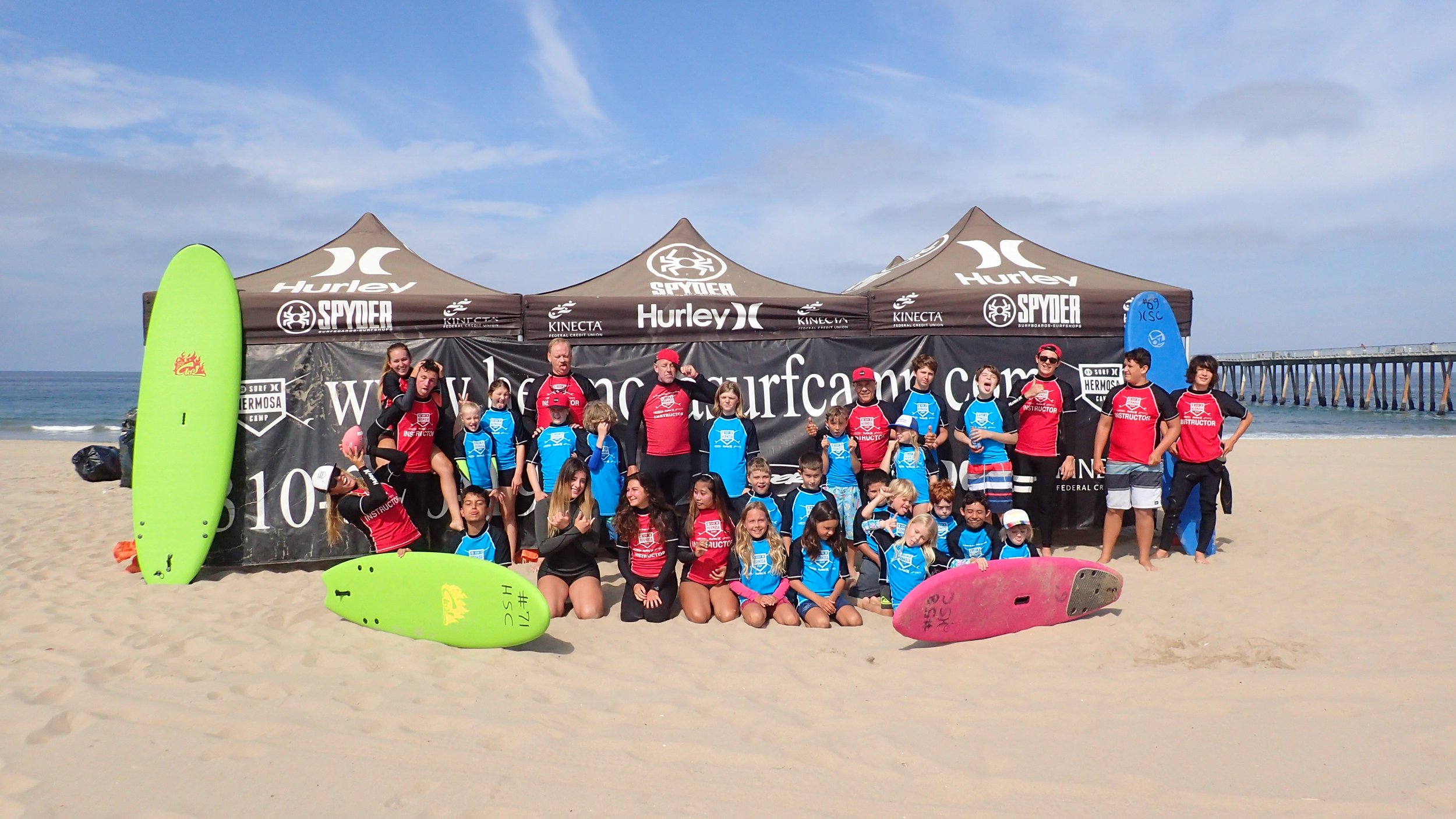 Group of children and instructors posing on a sandy beach with surfboards, tents, and a pier in the background, during a surf camp event.