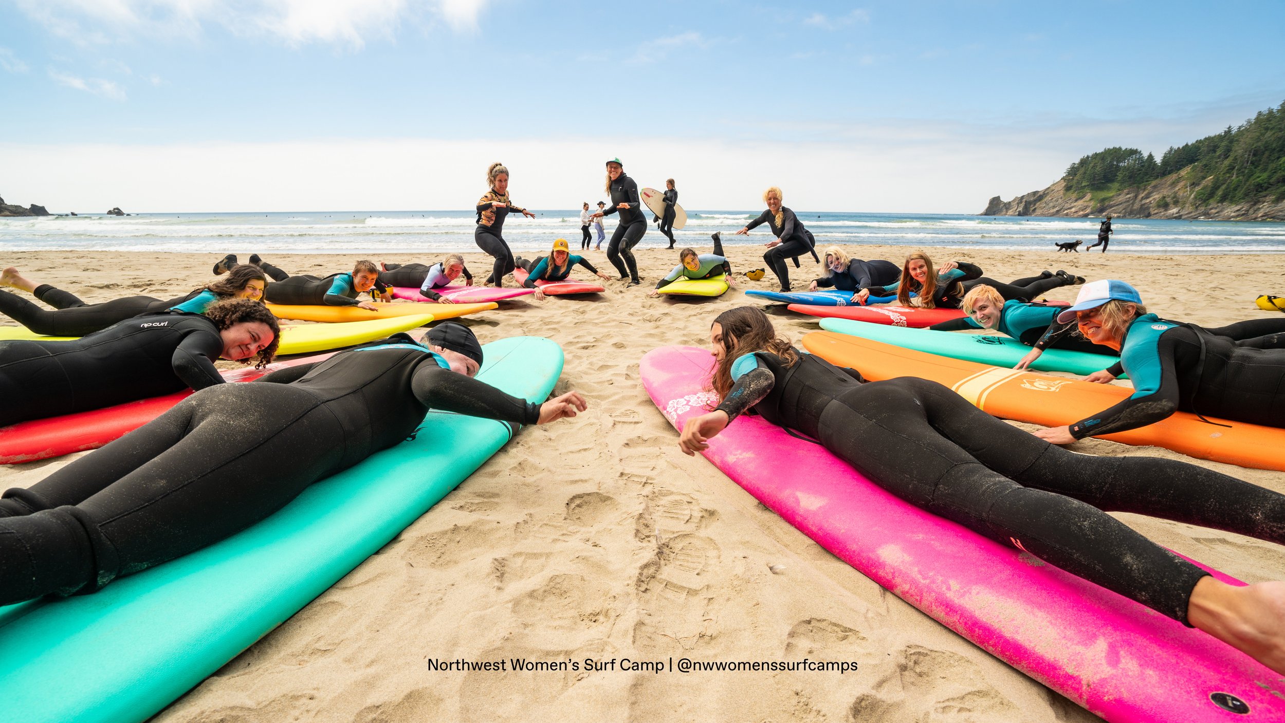 Group of women in wetsuits lying on surfboards on a sandy beach, participating in a surf camp, with some standing and holding surfboards near the ocean.
