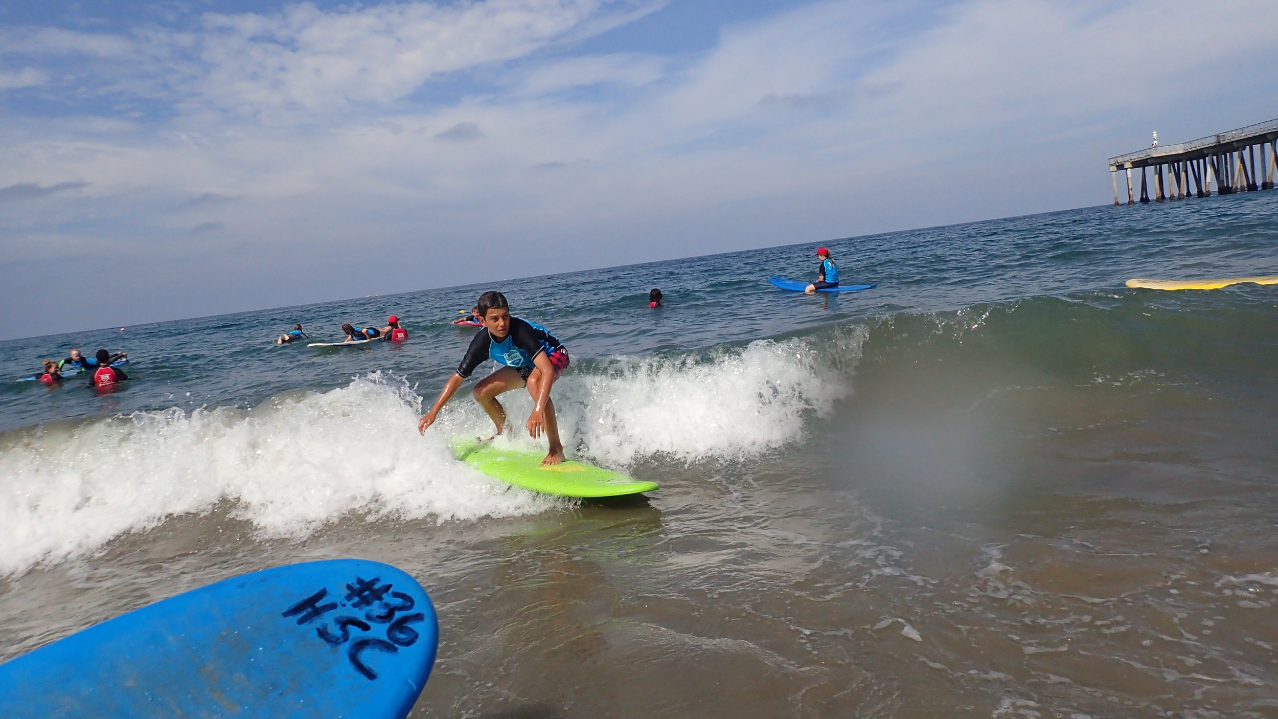 Boy surfing on a green surfboard at the beach with other surfers and a pier in the background.