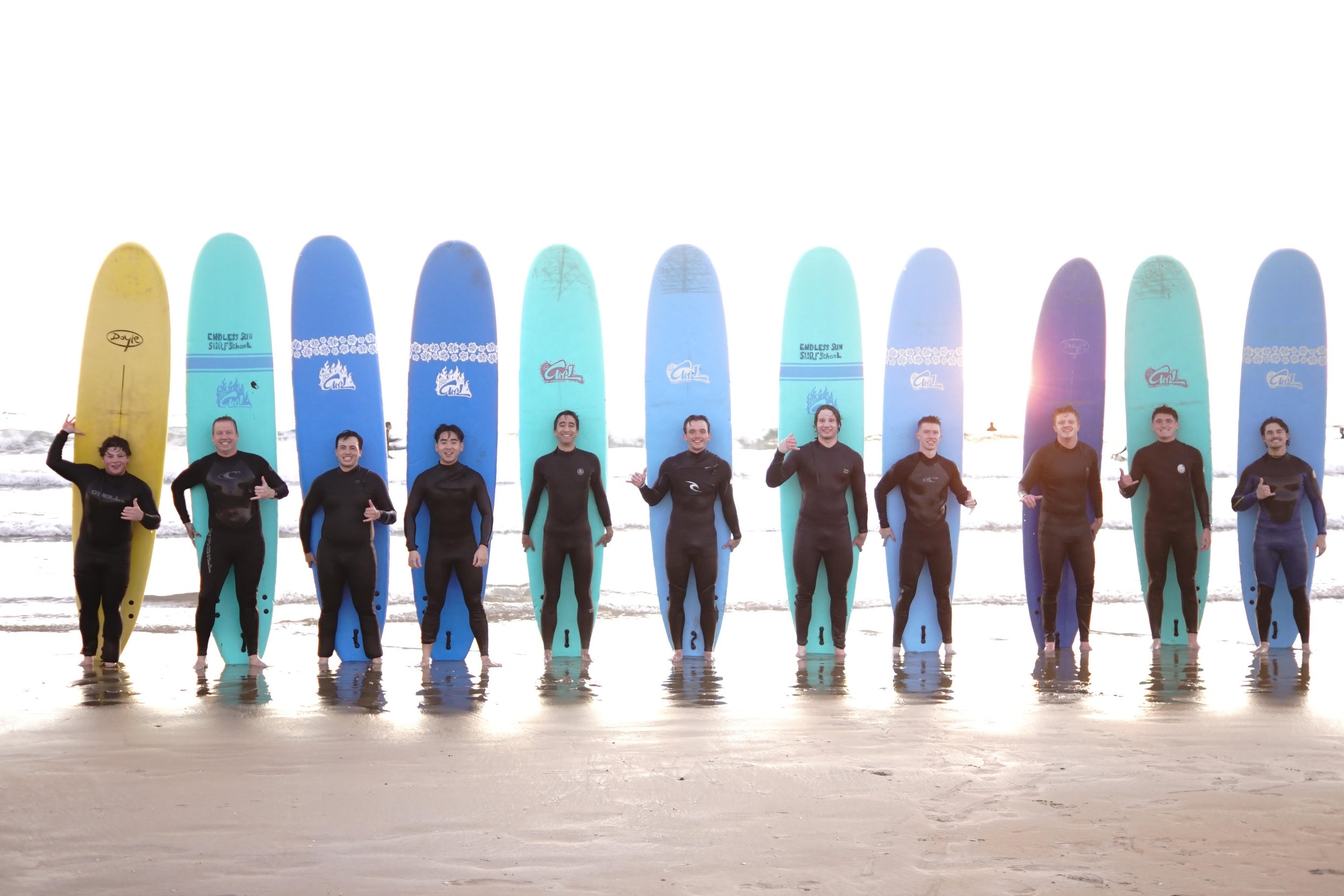 Group of eleven people in wetsuits standing in shallow water on the beach, each holding a surfboard upright behind them, with the ocean and a sunset in the background.