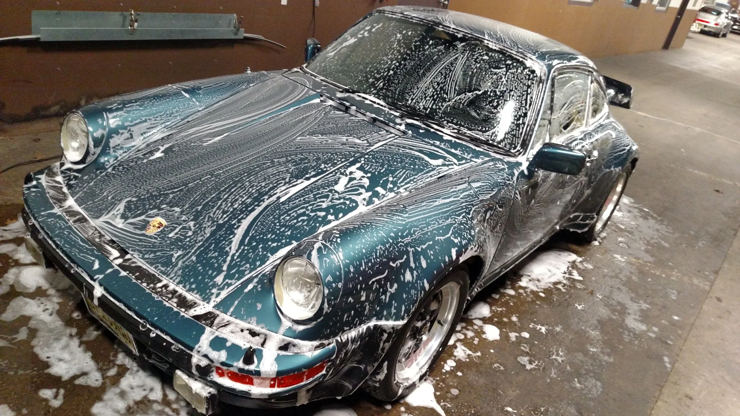 A blue vintage Porsche 911 sports car being washed, covered in soap suds with soap running down its hood and windshield in a parking garage.