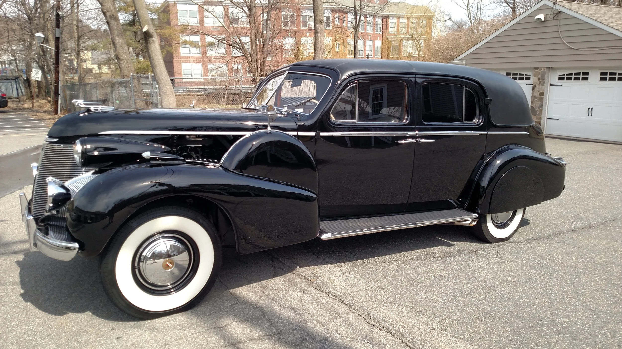 A vintage black sedan with white-wall tires parked on a paved lot with trees and residential buildings in the background.
