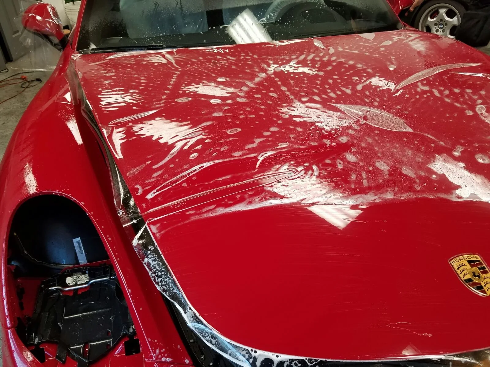 Red Porsche sports car with soap and water suds on the hood, in a garage or workshop setting.