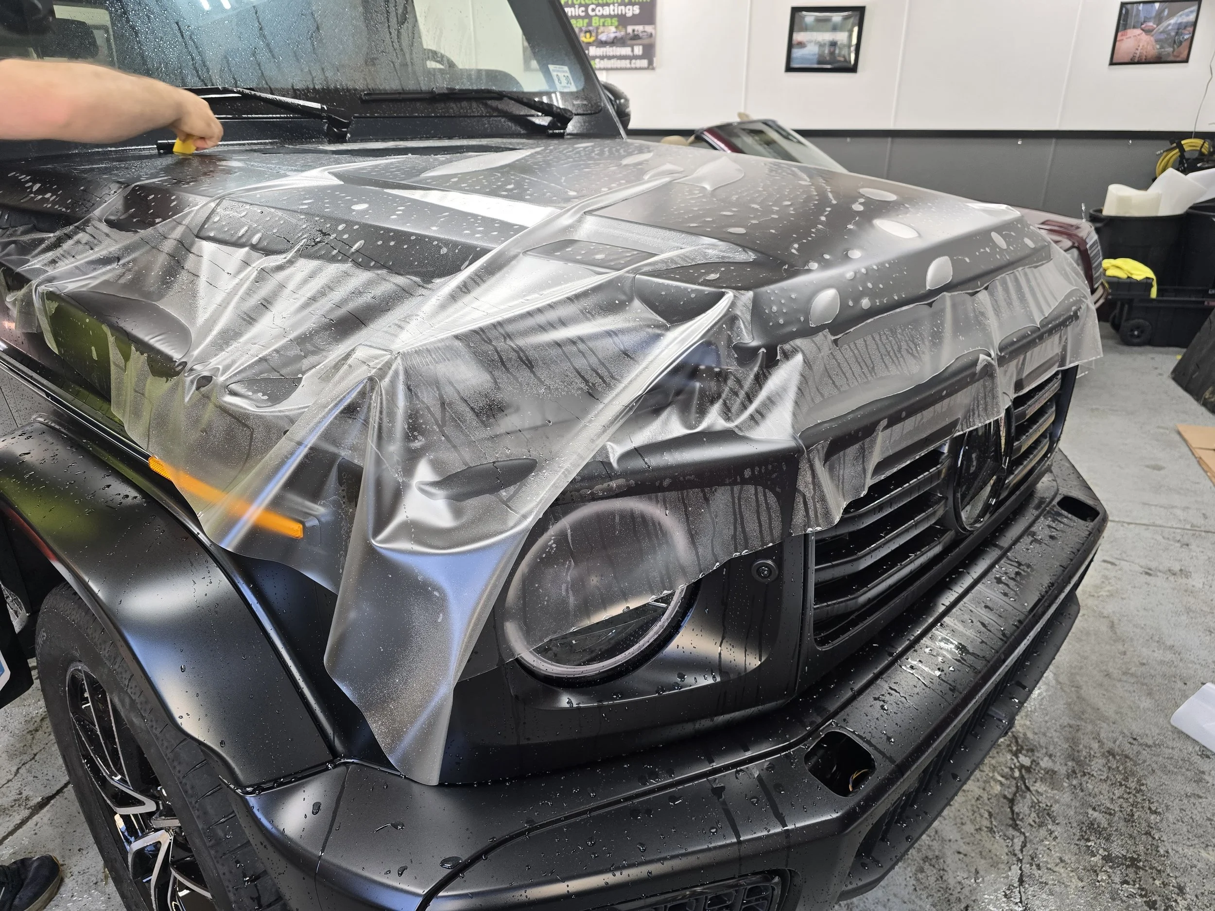 A black car being wrapped with a protective film, with a person applying the film while inside a garage or workshop.