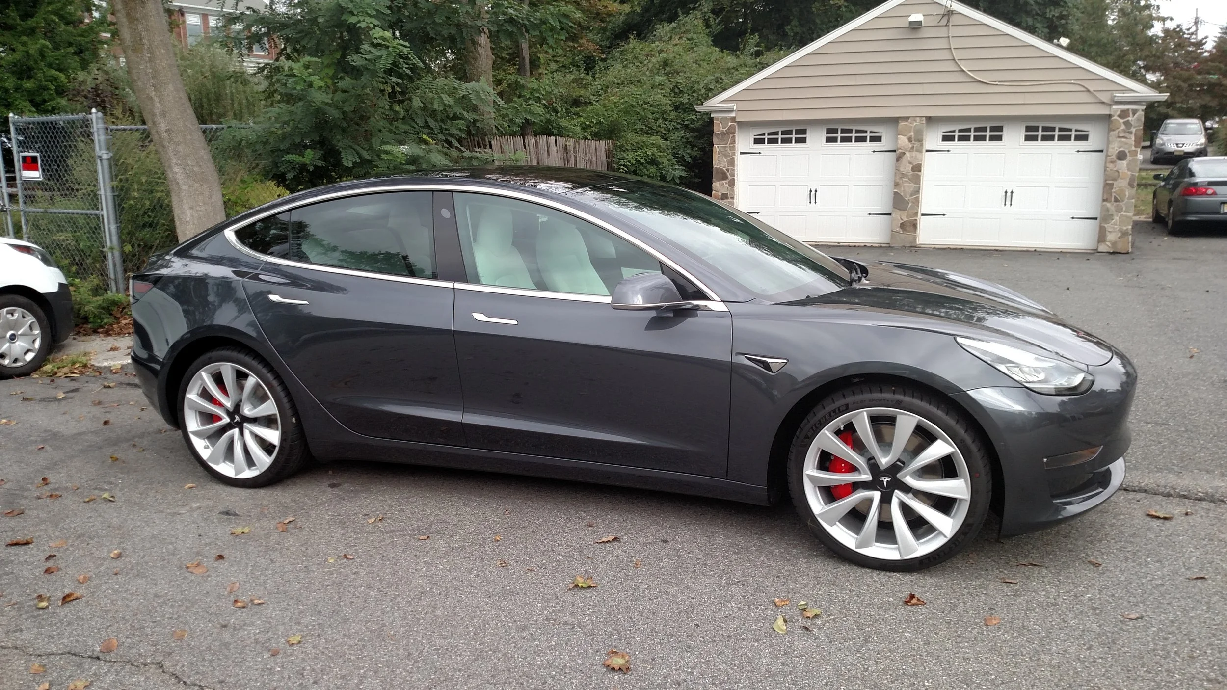 A black Tesla Model S parked in a residential driveway with a garage in the background, surrounded by trees and neighboring cars.