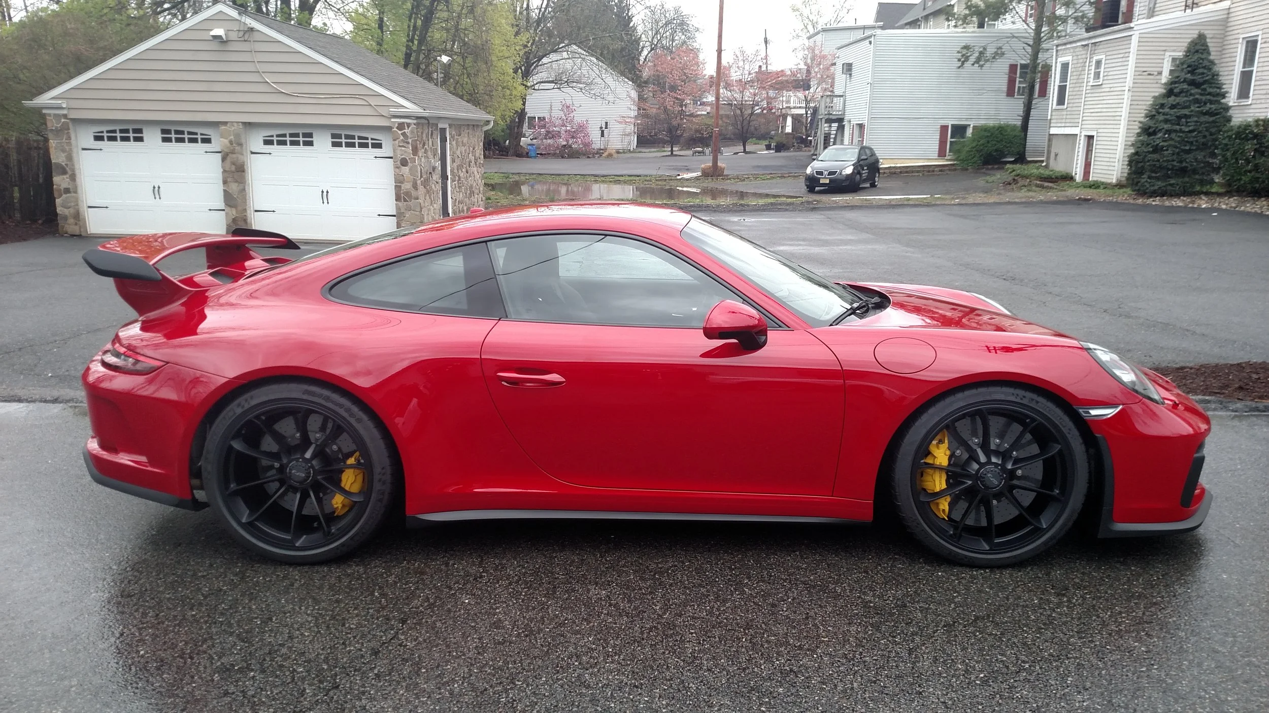 Red sports car with black wheels and yellow brake calipers parked on a wet asphalt driveway, houses and trees in the background.