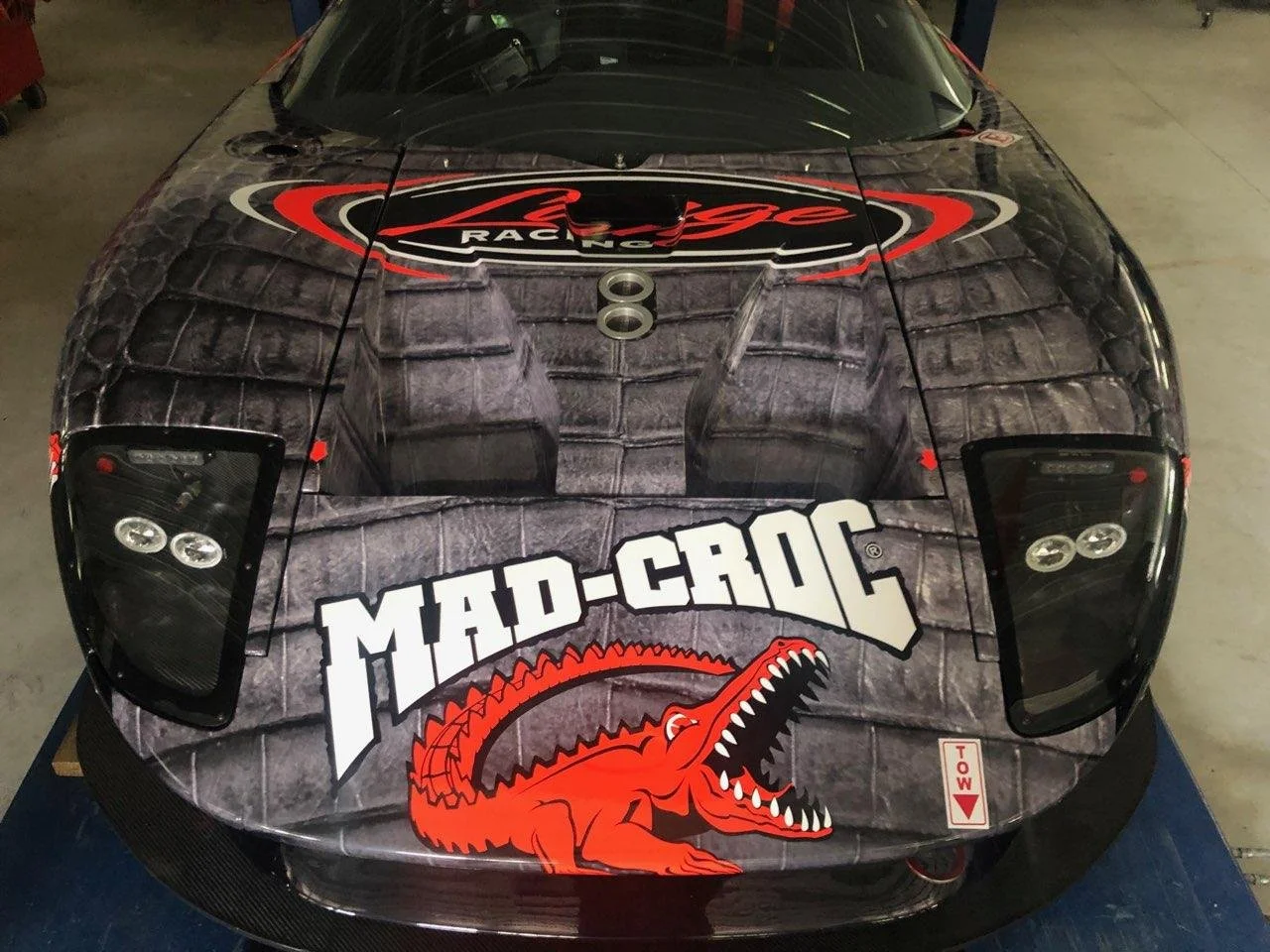 Close-up of a race car with Mad Croc Racing branding and a large red crocodile logo. The car has a dark, textured hood and distinctive headlights.
