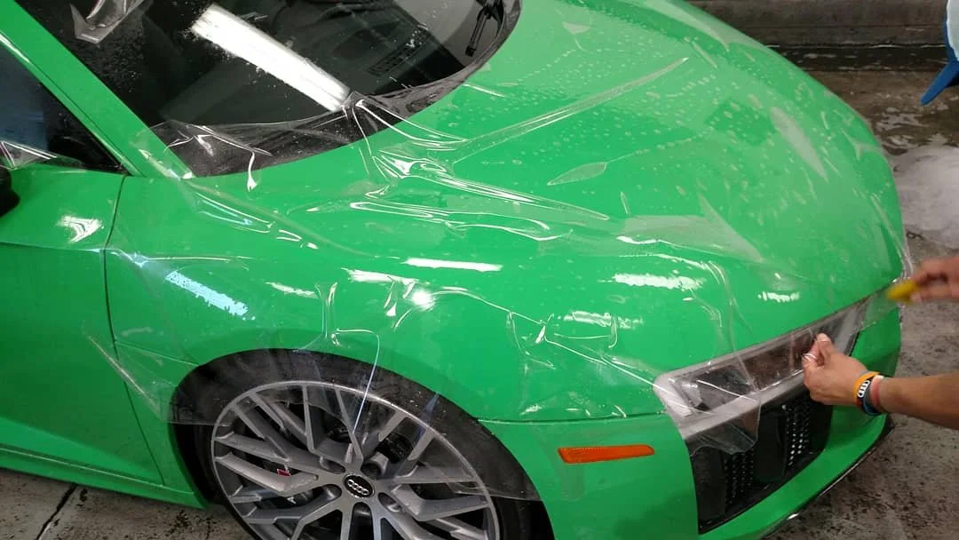 A person applying clear protective film to a bright green sports car in a garage or workshop.