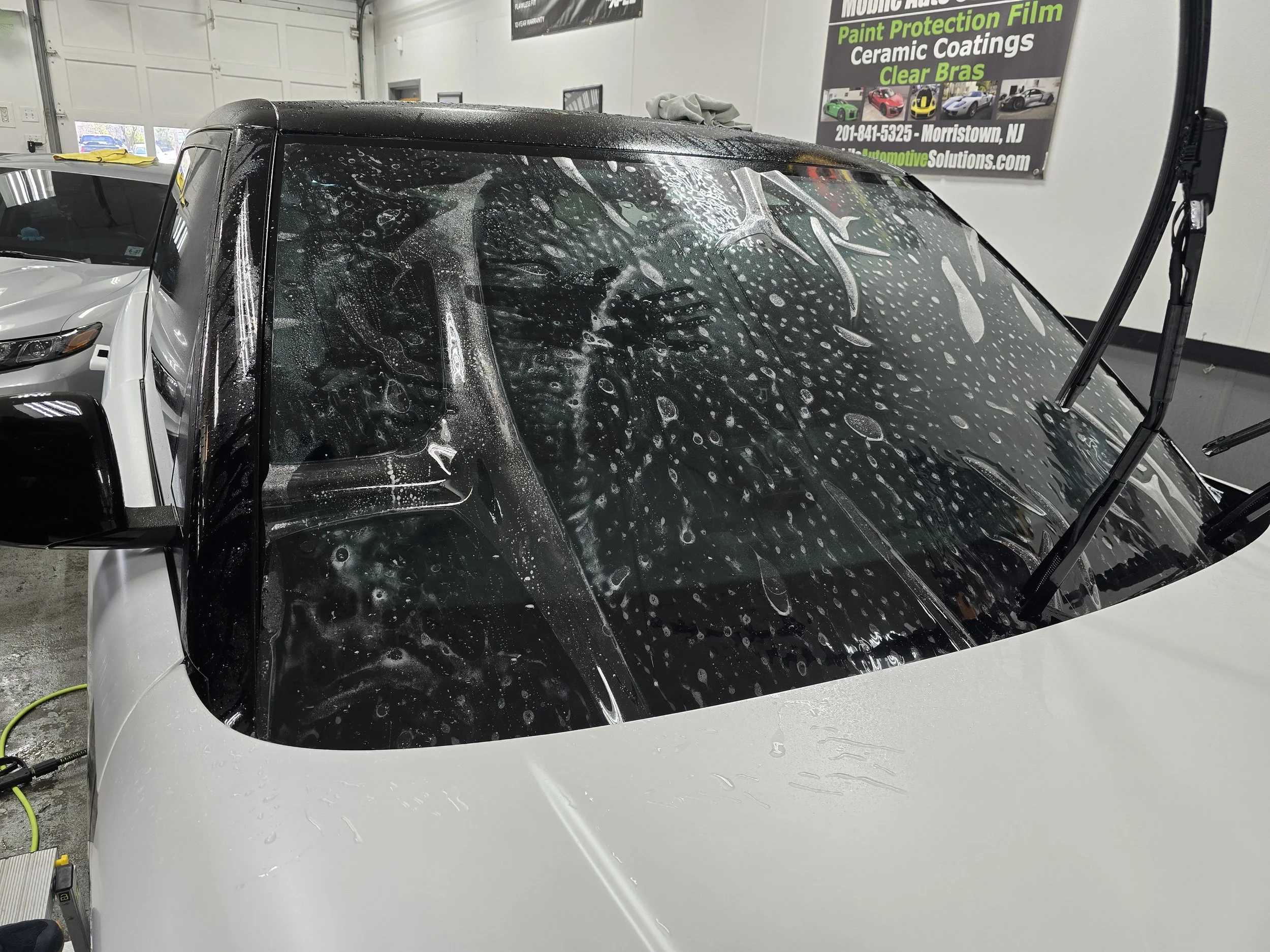 A white sports car with a black windshield and roof being washed or detailed inside a garage. Water soap and bubbles are visible on the windshield, and there is a side mirror on the left. A sign in the background advertises automotive services.