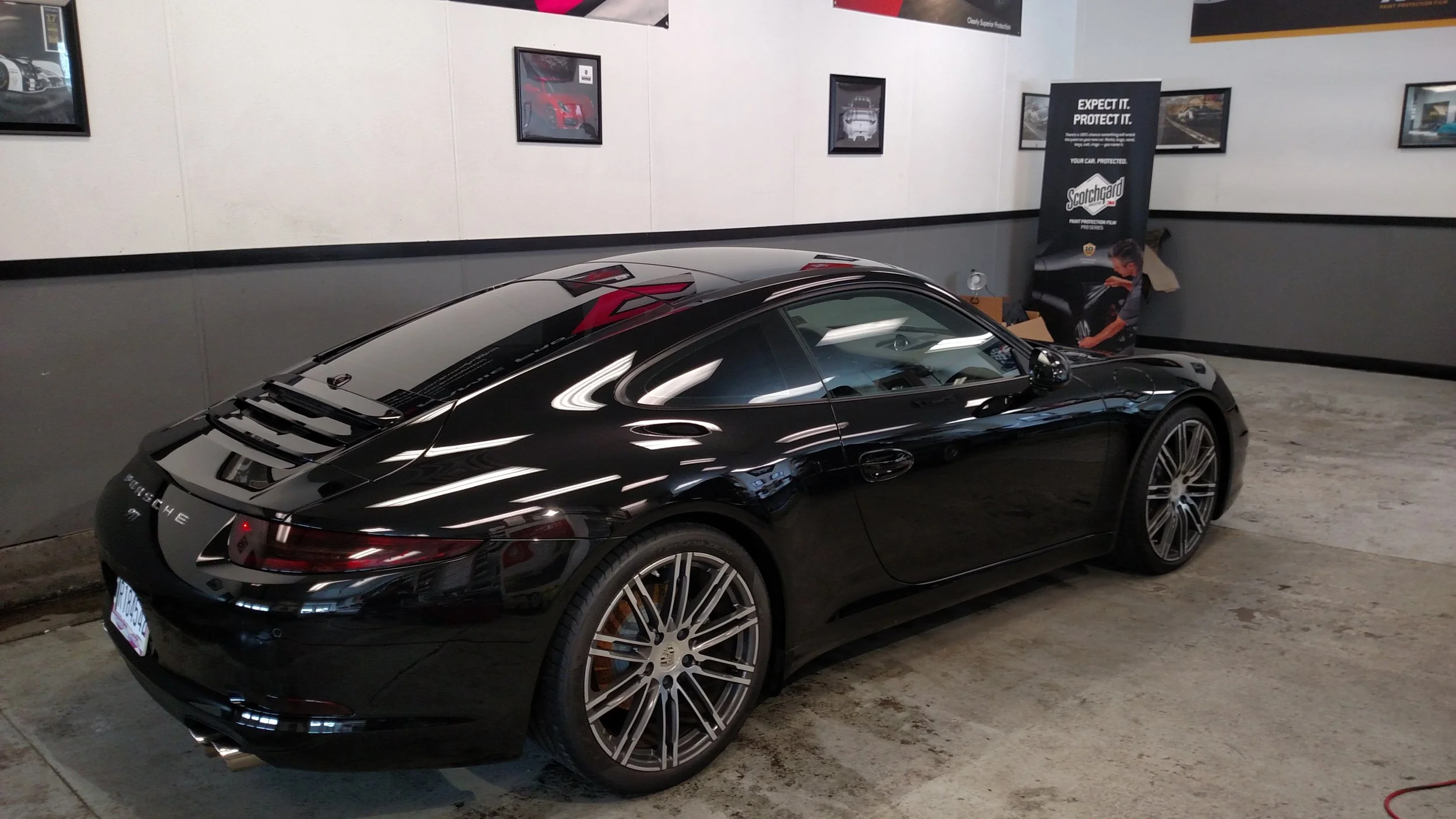 Black Porsche convertible parked indoors with posters on the wall and a person working on tools in the background.