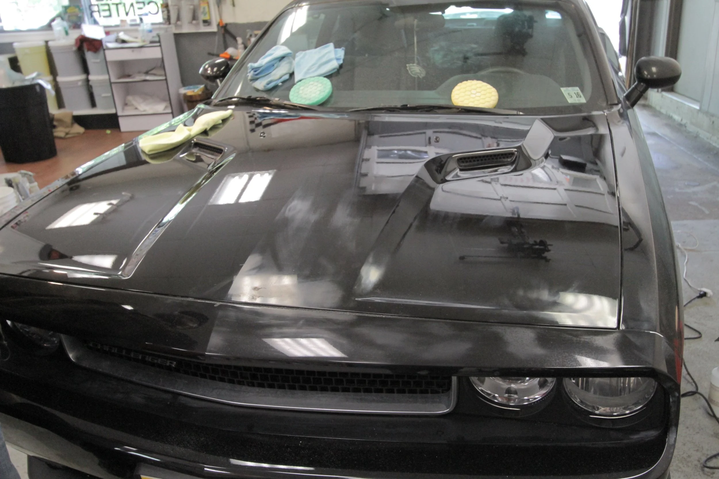 A black Dodge Challenger in a garage undergoing detailing, with polishing pads, towels, and polishing compounds placed on the hood and windshield.