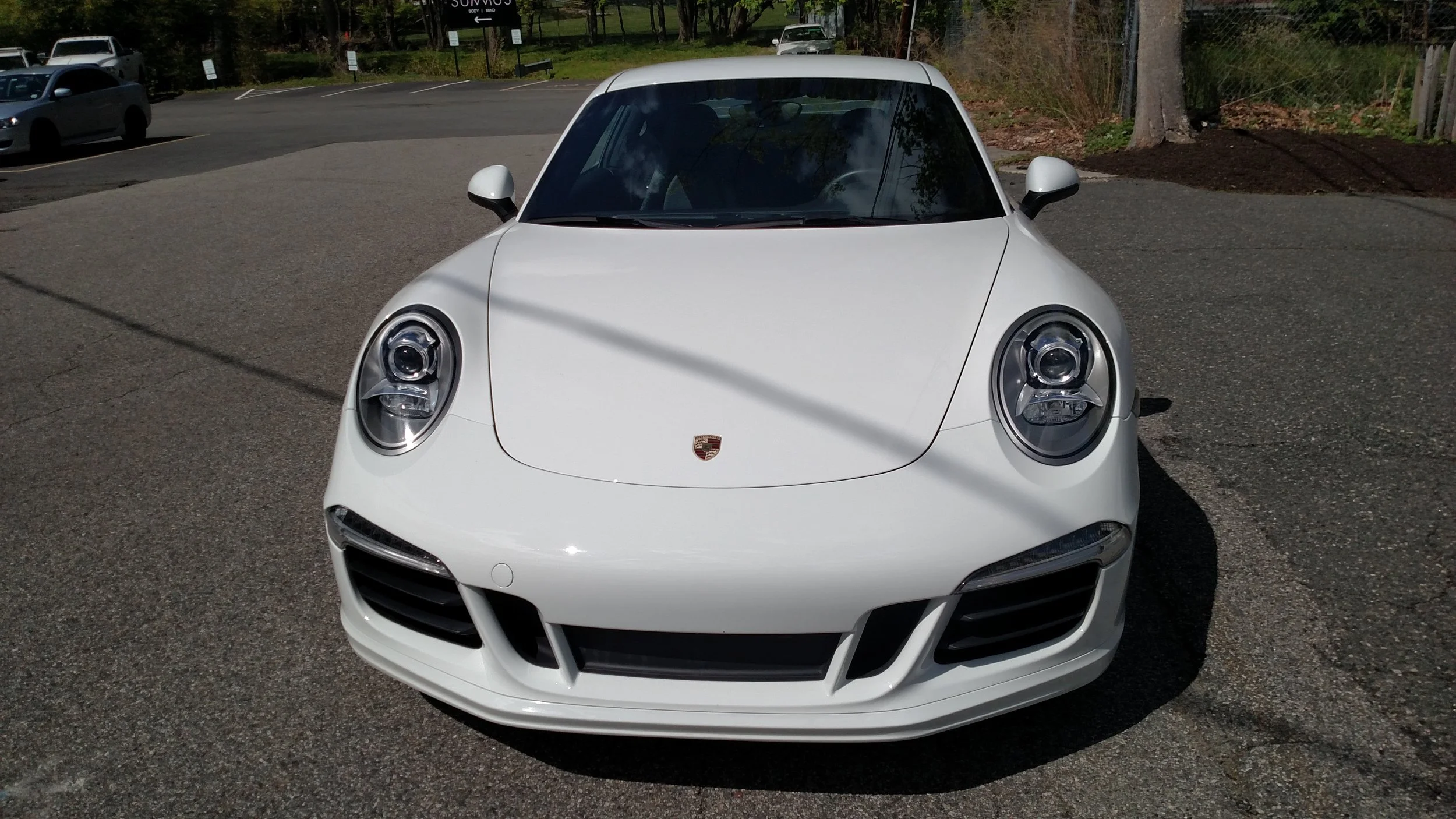 Front view of a white Porsche sports car parked in an outdoor lot, with trees and other parked cars in the background.