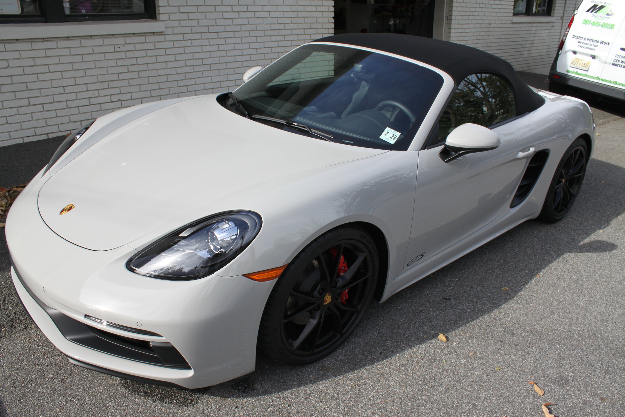 A white Porsche sports car with black wheels and a black convertible top parked on a street in front of a white brick building.