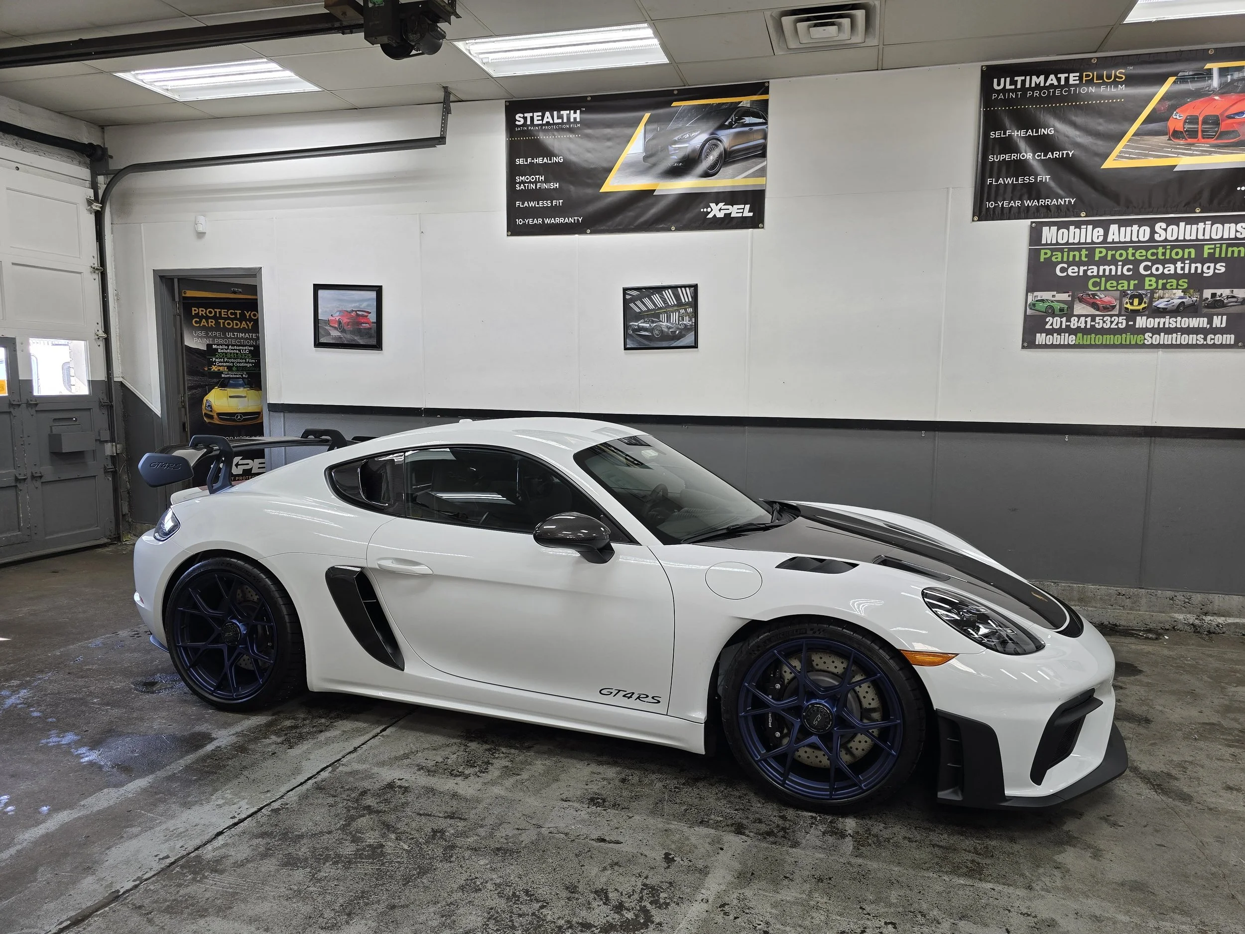 White sports car with black accents in a garage setting, featuring a large rear wing and custom wheels.