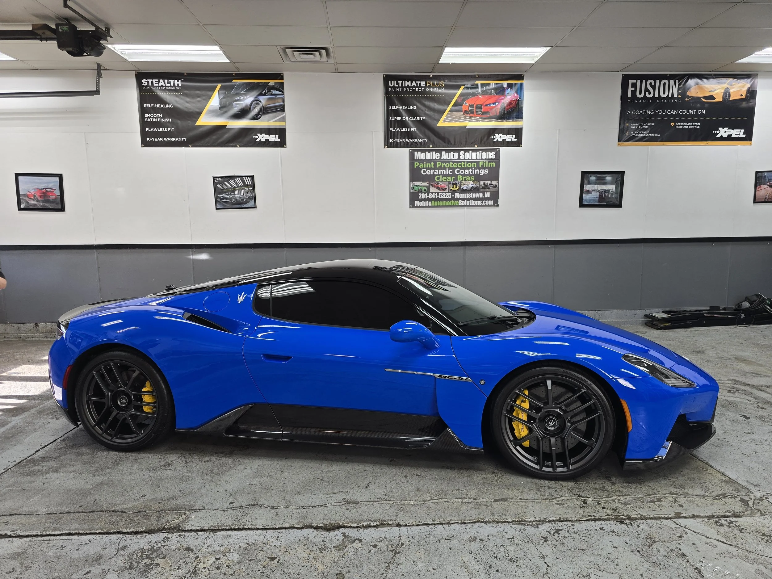 A blue sports car with black wheels and yellow brake calipers parked indoors near a wall with advertisements and framed pictures.