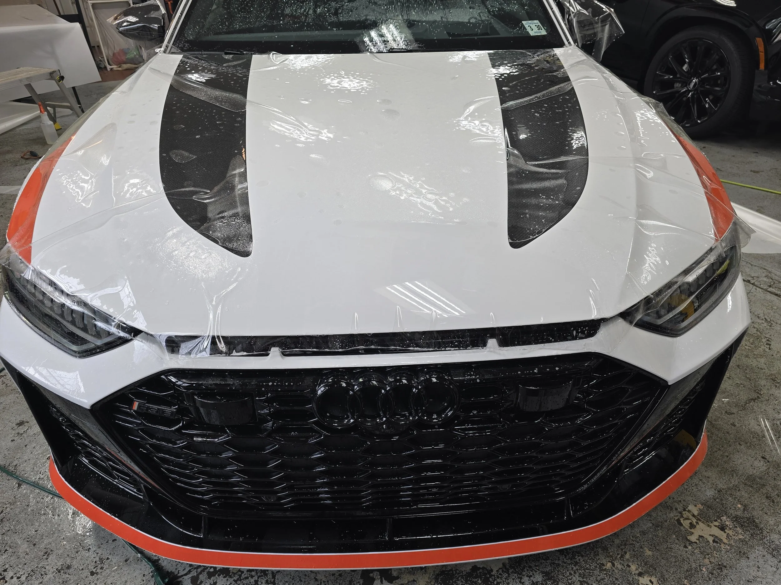 A white sports car with black and orange accents being wrapped with clear plastic film.